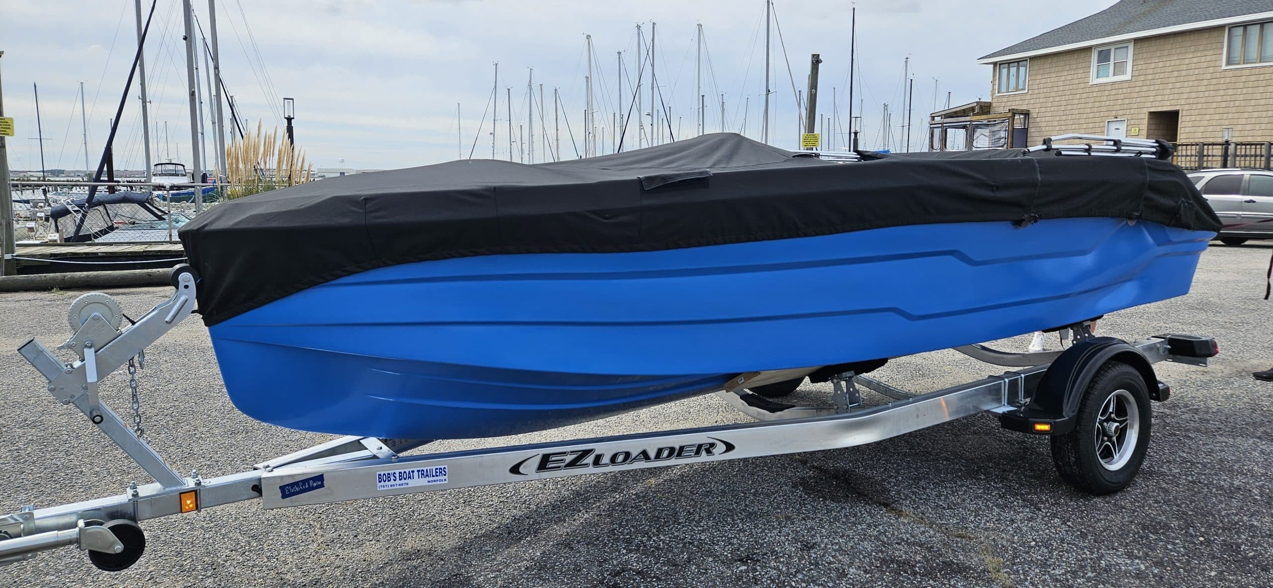 Blue Phantom powerboat covered on a trailer at a marina, with sailboats and waterfront buildings in the background.