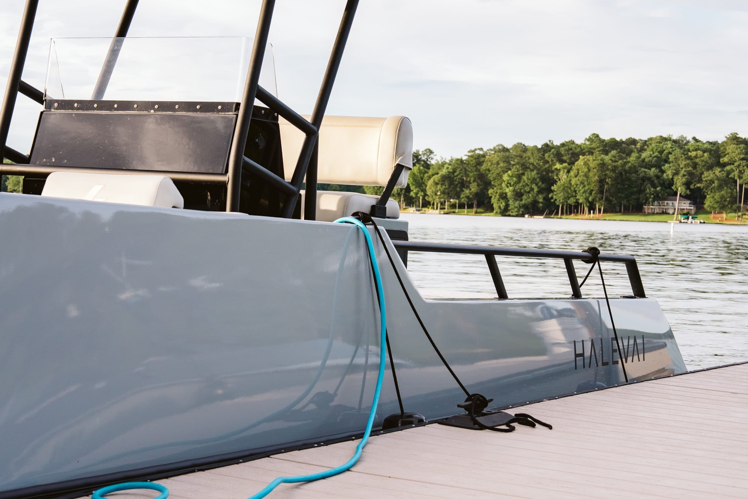Halevai electric boat hull with blue charging cable on a dock beside a tree-lined lake.
