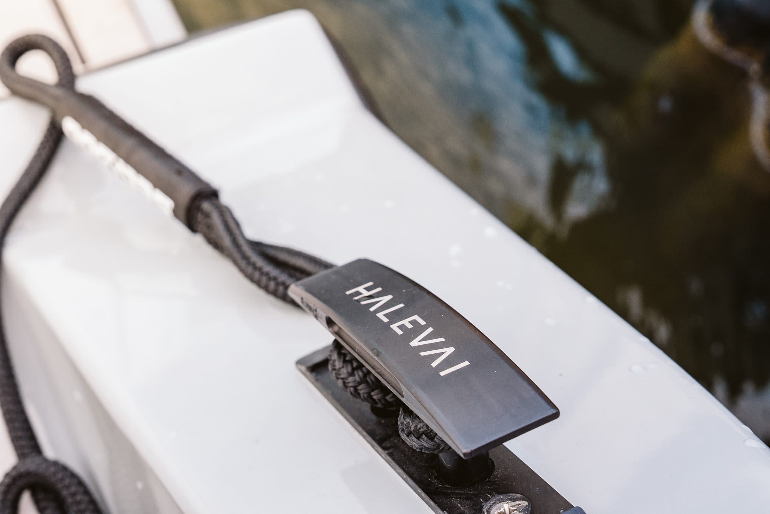 Close-up of a Halevai-branded black cleat component and dock line of the white deck of a boat beside the water.