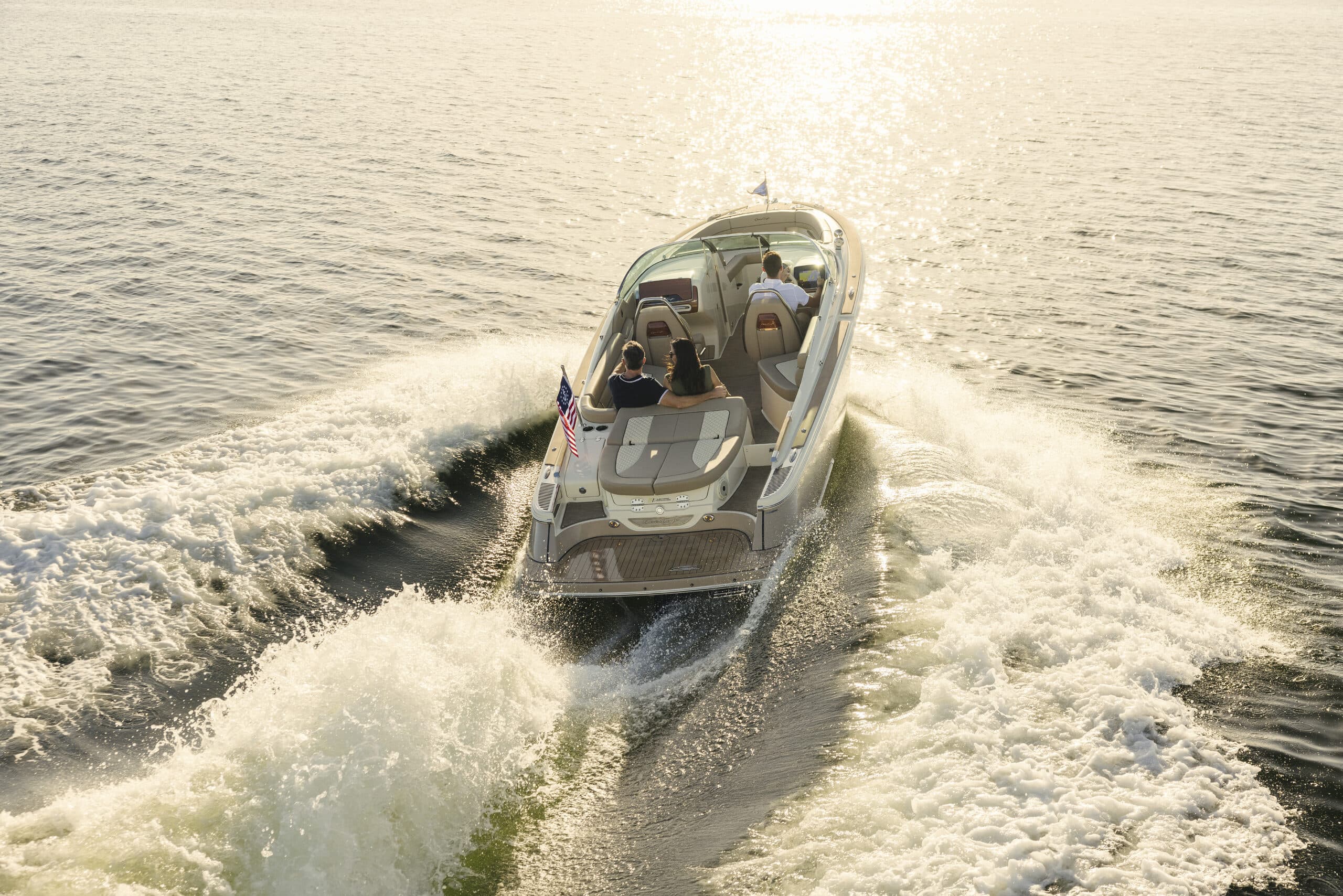 Chris-Craft Launch 25 GTe with three people on board, speeding across sunlit water, viewed from above with wake spreading behind the boat.