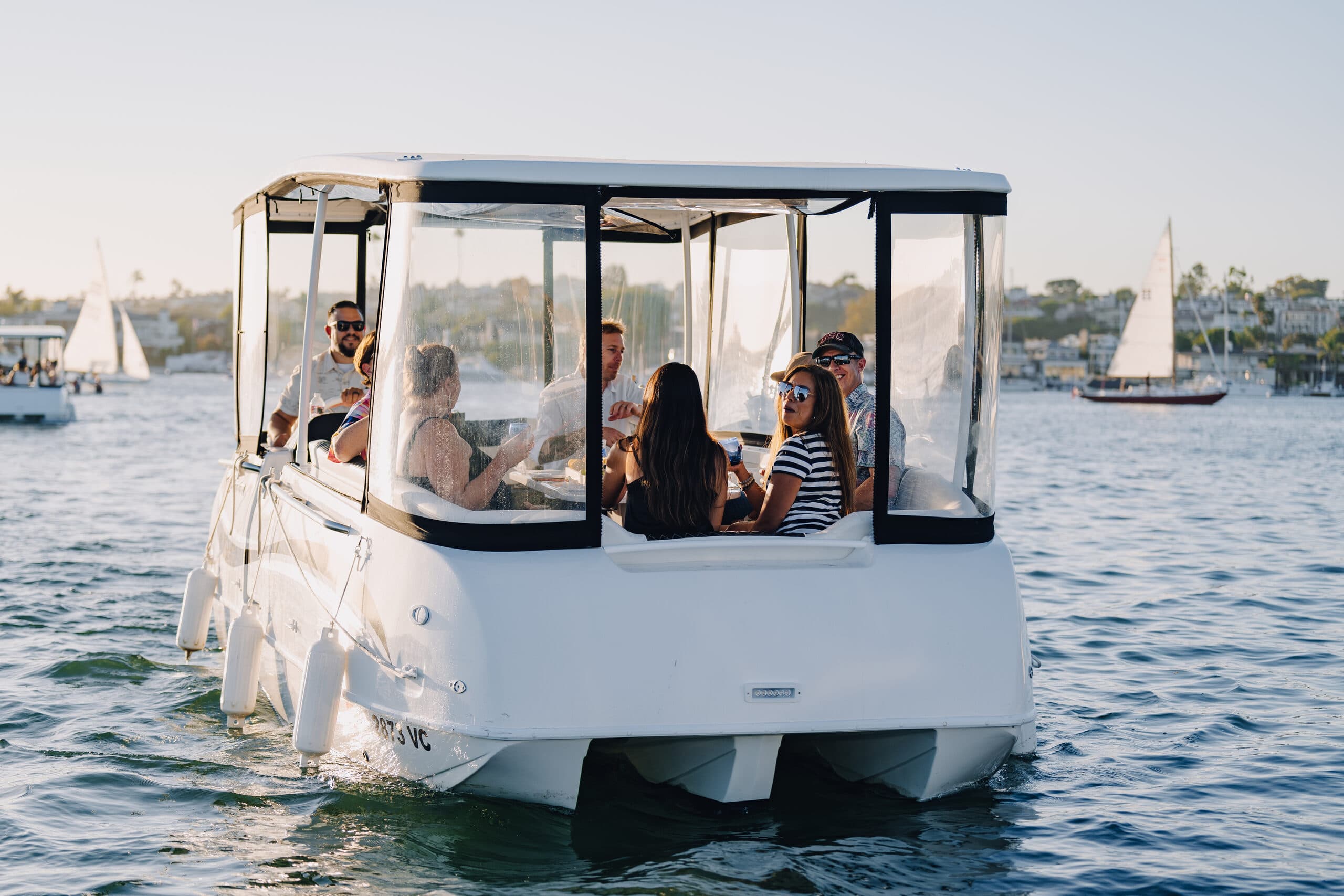 From behind ElectraCraft 182TR electric boat carrying seated passengers on calm water, with a canopy, clear enclosure panels, and sailboats in the background