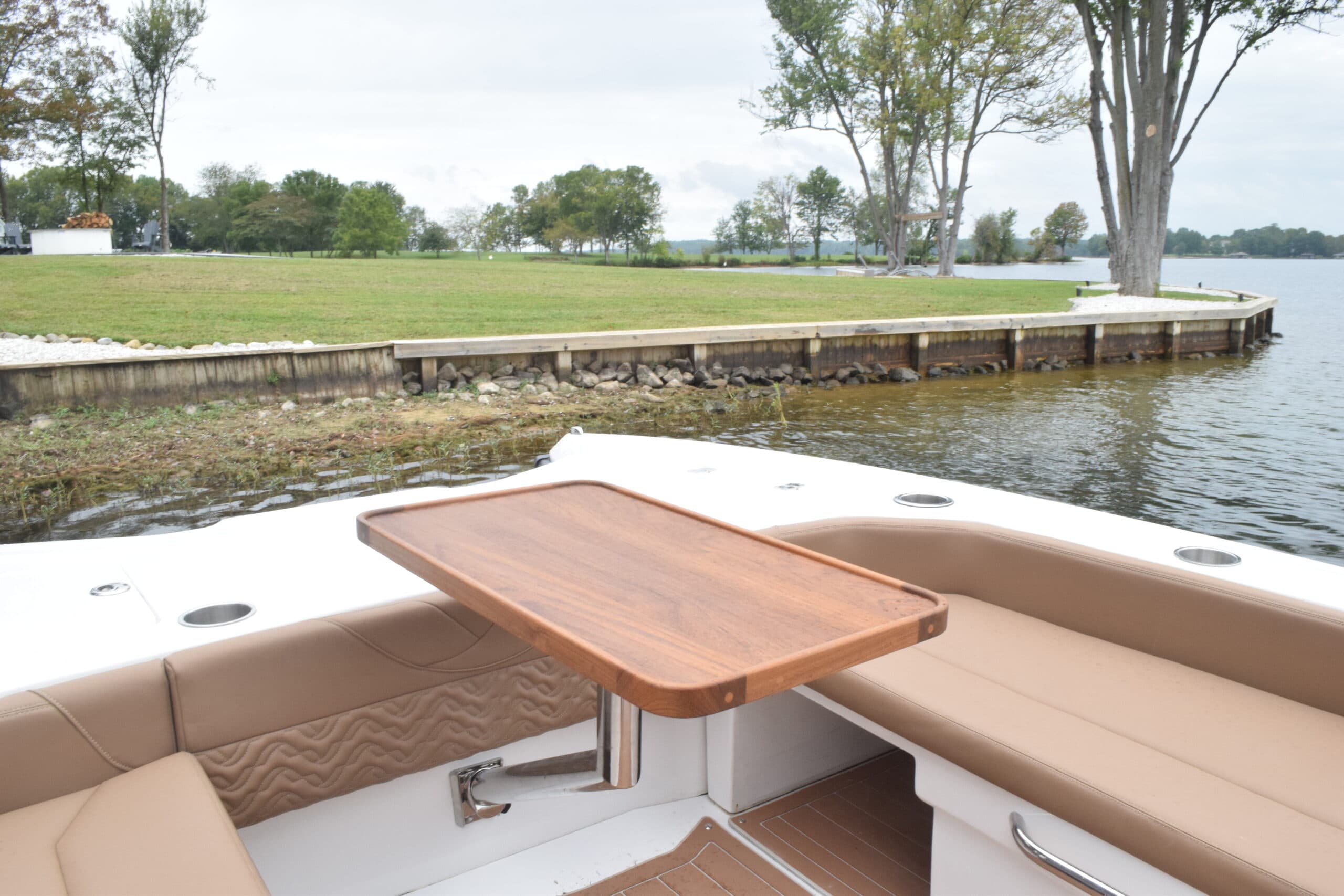 Volare Artemis 23 electric boat cockpit with wood table, tan seating, and shoreline visible beyond the stern.