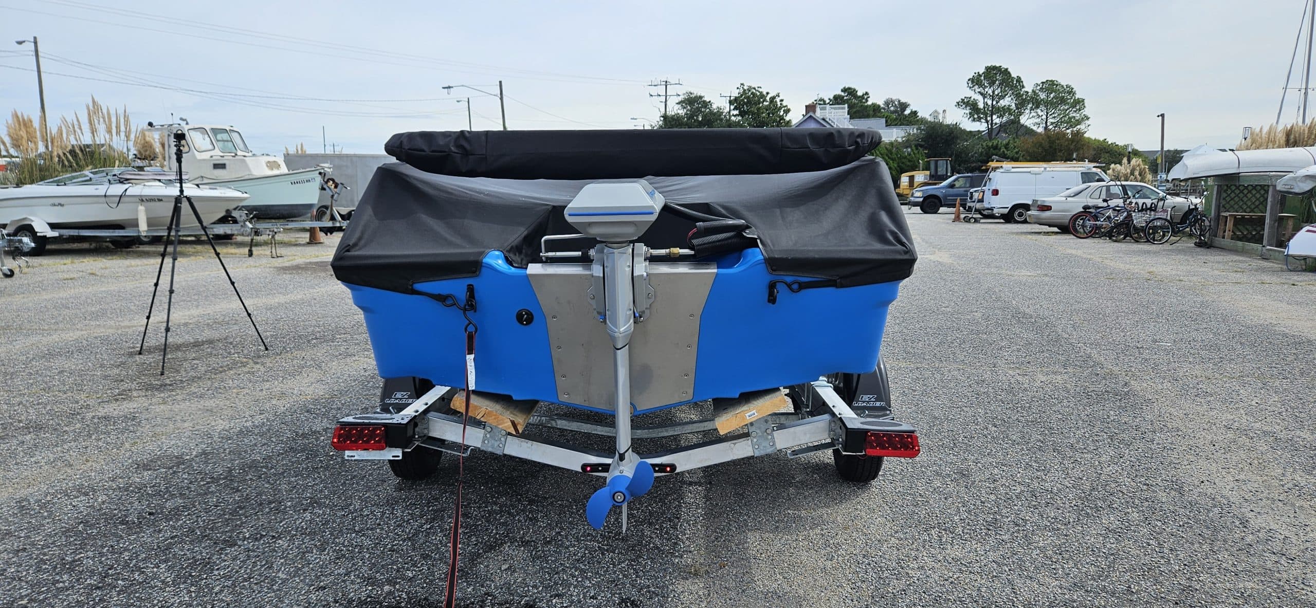 Rear view of a blue Vision Marine Phantom electric boat on a trailer in a gravel marina lot, with a covered cockpit and outboard motor.