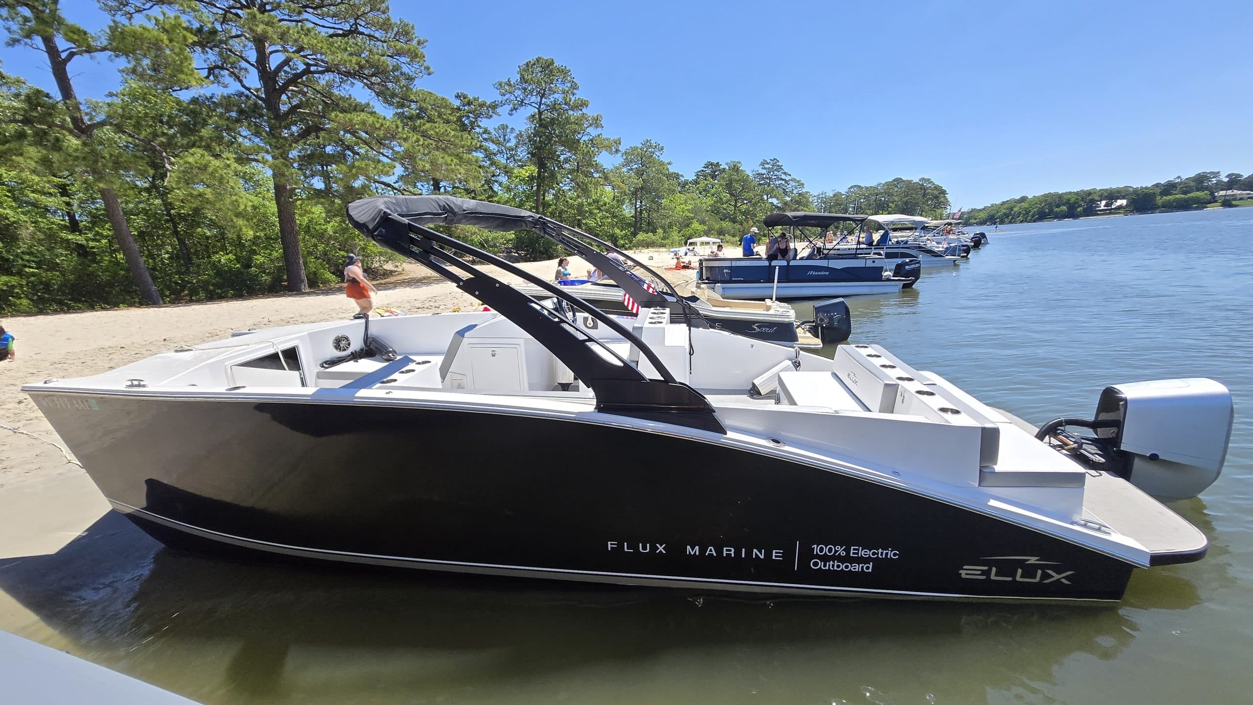 GFX Stealth electric catamaran style beached at a lakeside, with an open deck, black canopy frame, and outboard motor visible.