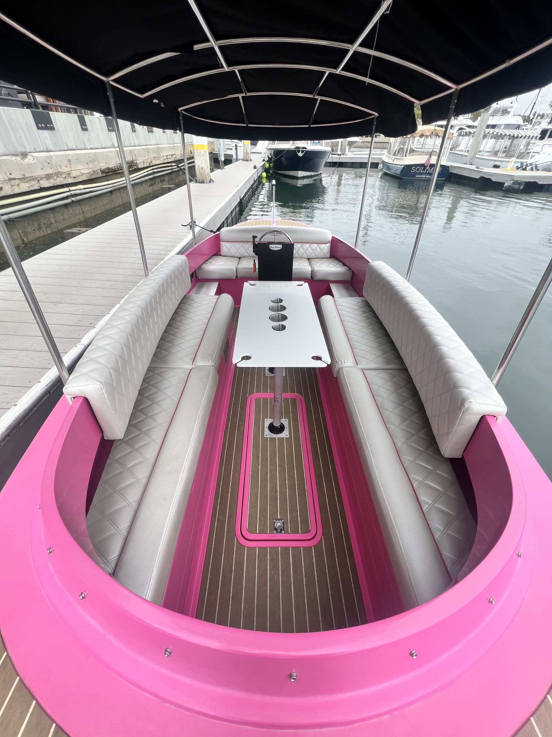 2024 Vision Marine Fantail 217
Interior of a pink-and-white Vision Marine Fantail with white quilted cushioned seating, central table, canopy, and marina dock visible.