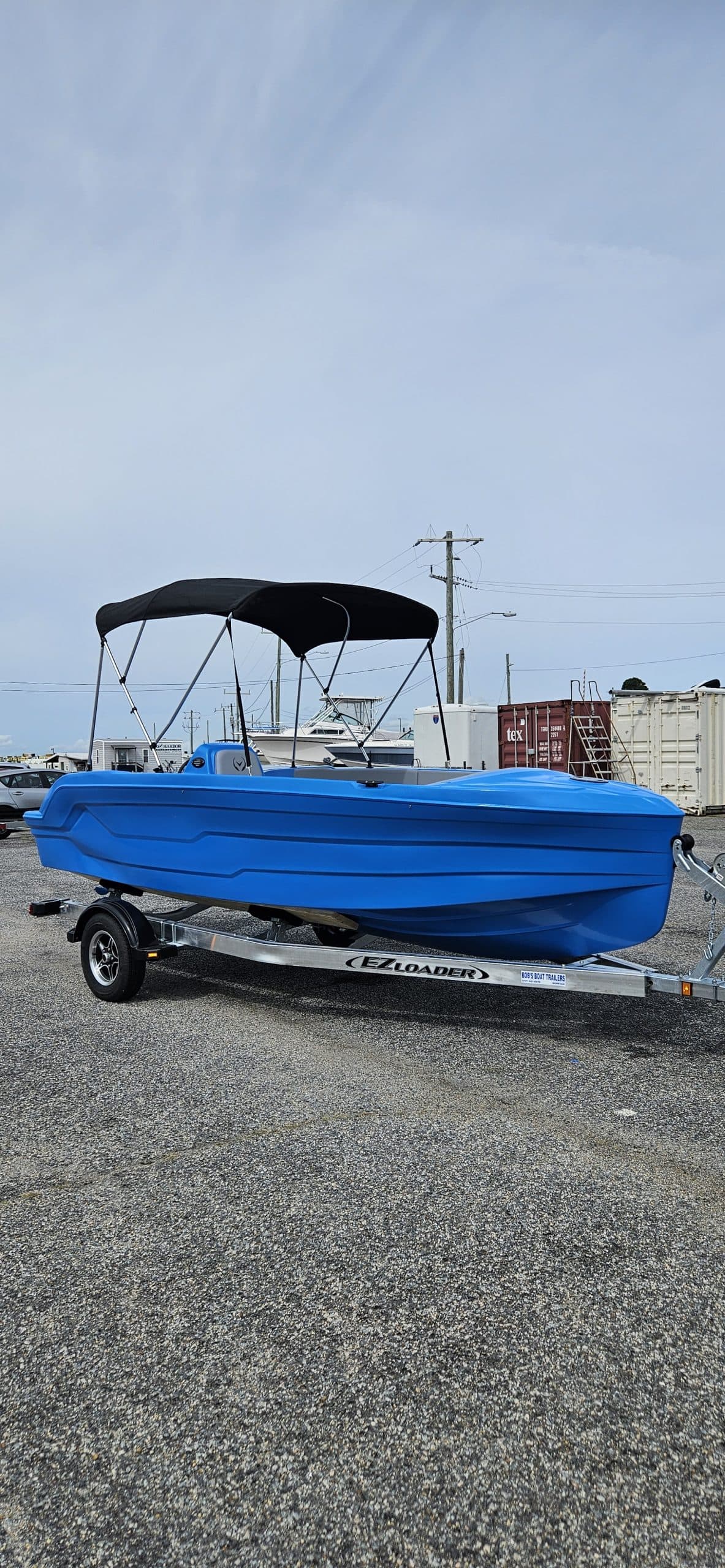 Blue Vision Marine Phantom boat on a trailer outdoors, with a canopy and industrial storage containers in the background.