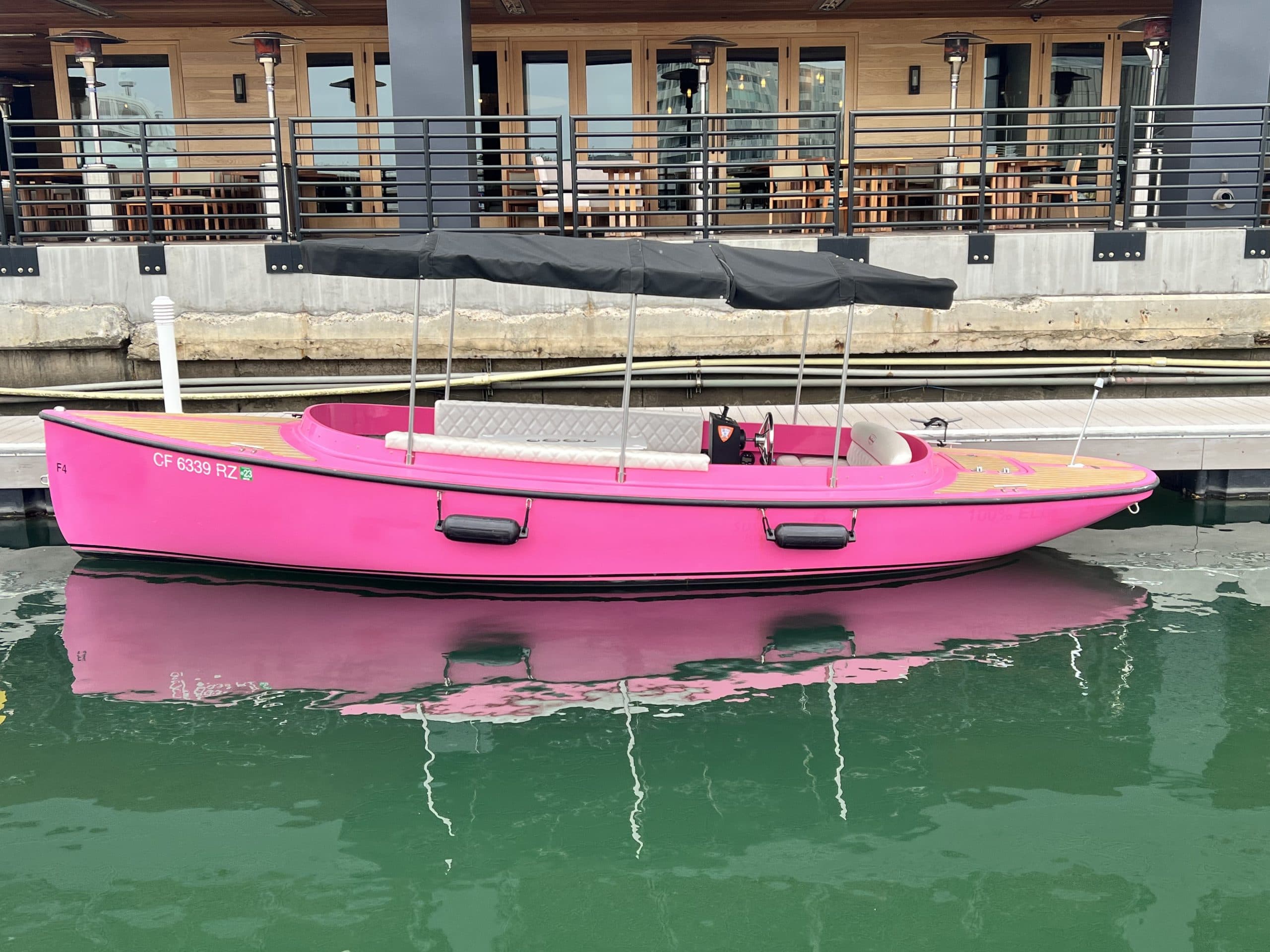 2024 Pink Vision Marine Fantail electric boat moored along a waterfront dock, reflected in calm green water.