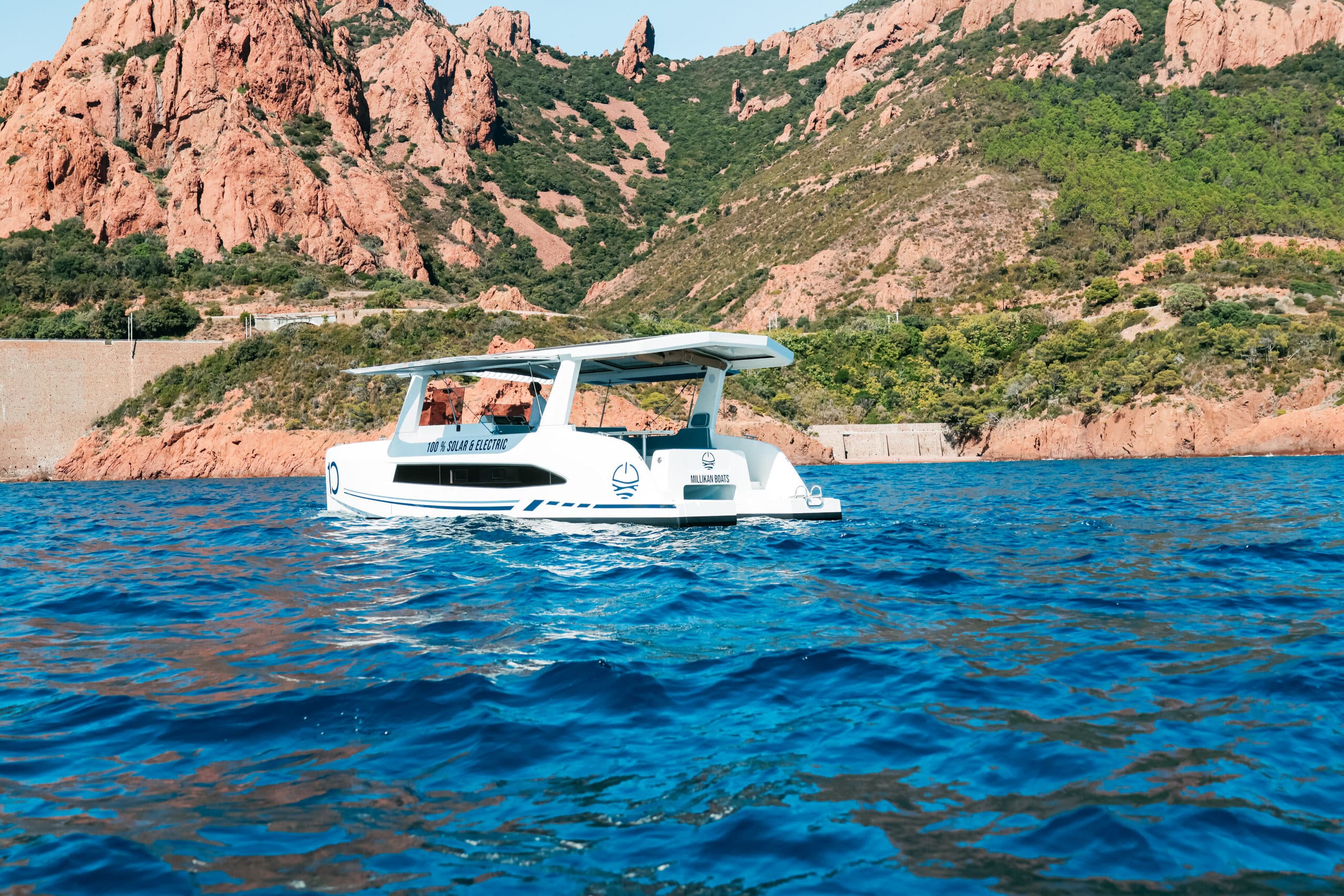 Millikan M10 boat viewed from the side on blue water, with rocky coastal mountains in the background