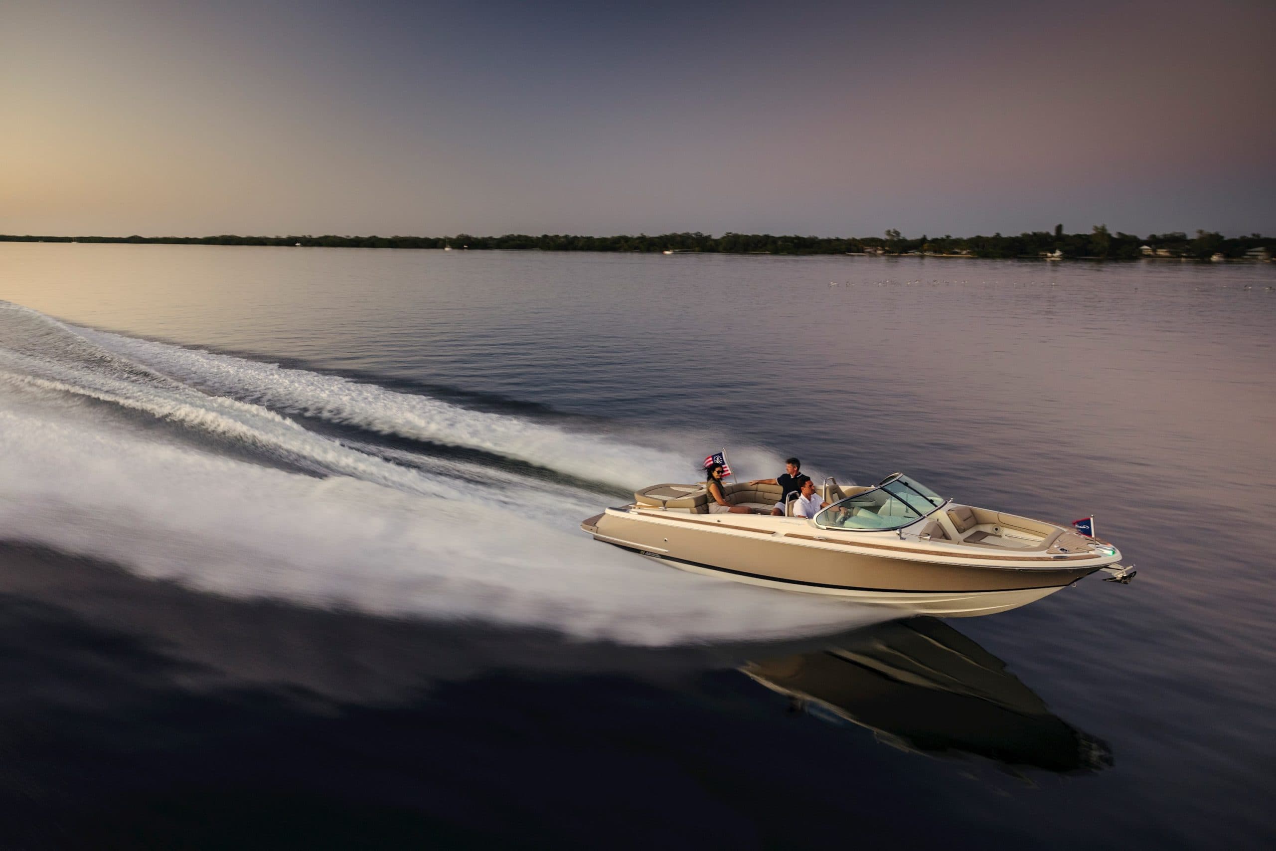 Chris-Craft boat bowrider speeding across calm water at dusk, leaving a wake with two people aboard.