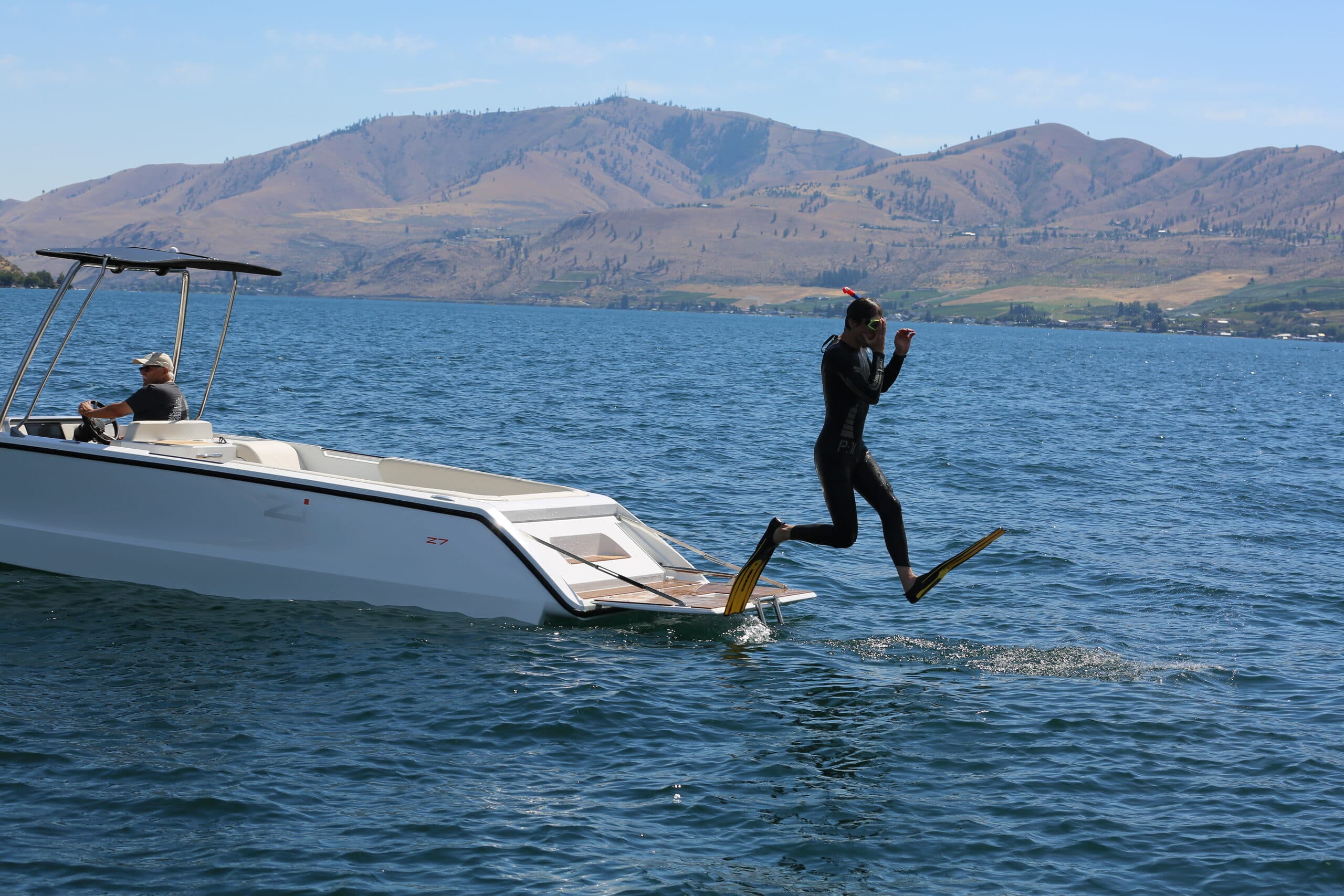 Scuba diver jumping off the back of swim platform on white boat (2026 Zin Boats Z7). Behind him are mountains