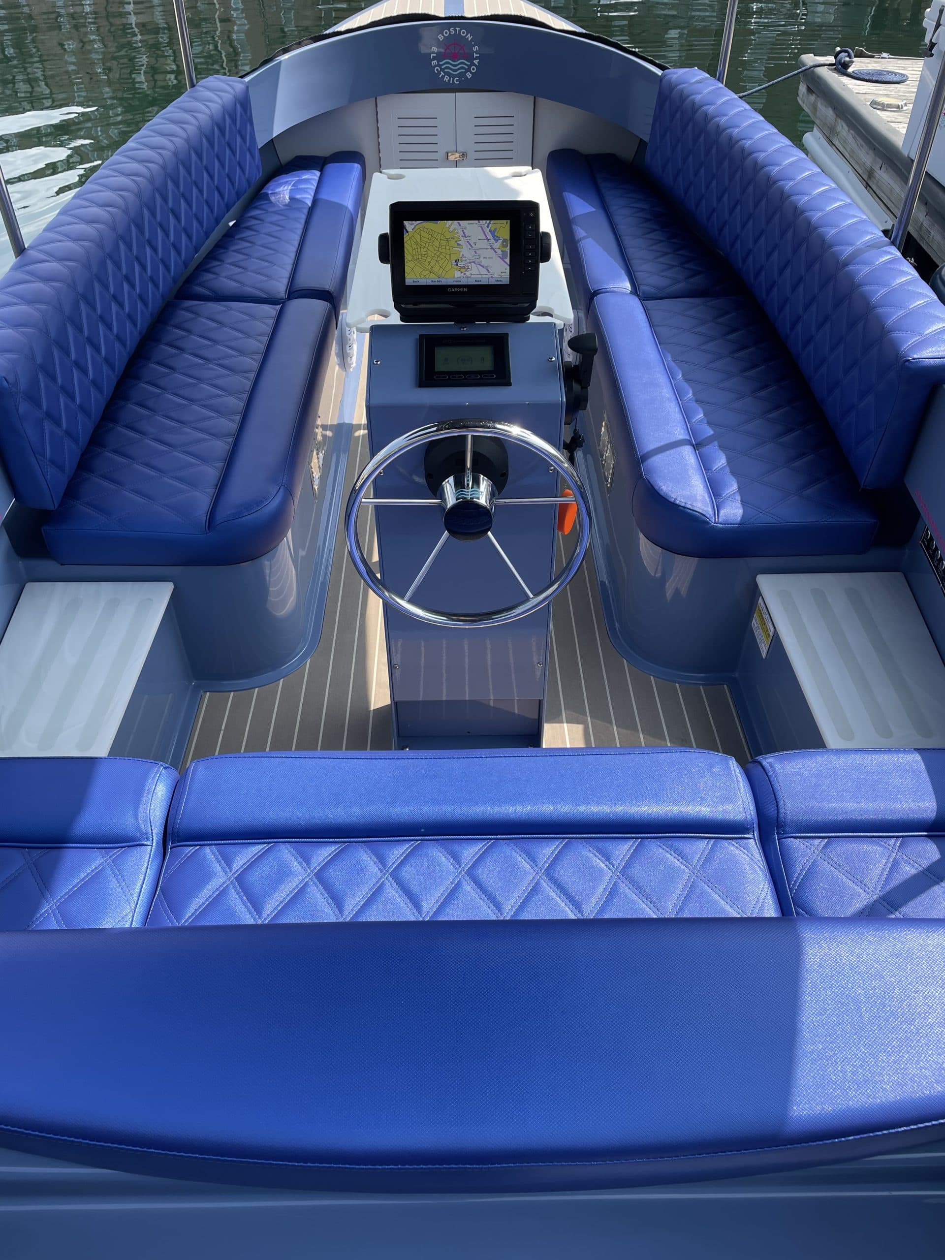 Vision Marine Fantail 217 blue-upholstered electric boat cockpit with wraparound seating, steering wheel, dashboard displays, and dockside water visible beyond the bow.