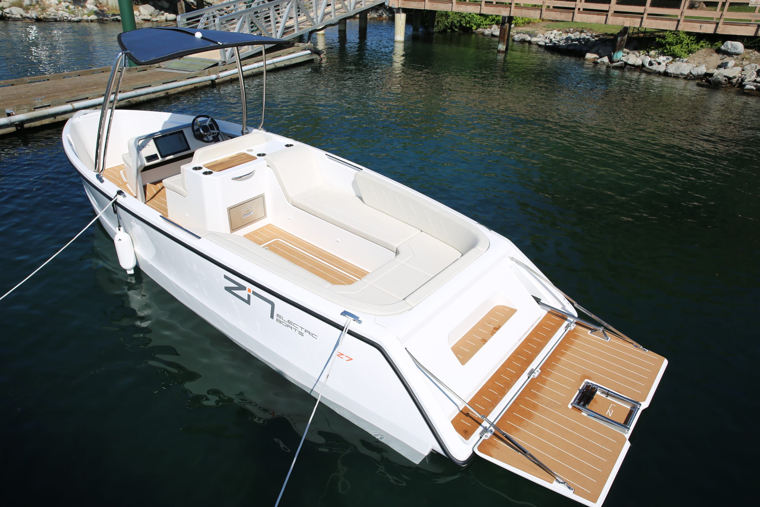 A sleek white motorboat with wooden deck details docked by a pier. It features cushioned seating, a steering console, and calm water surroundings.
