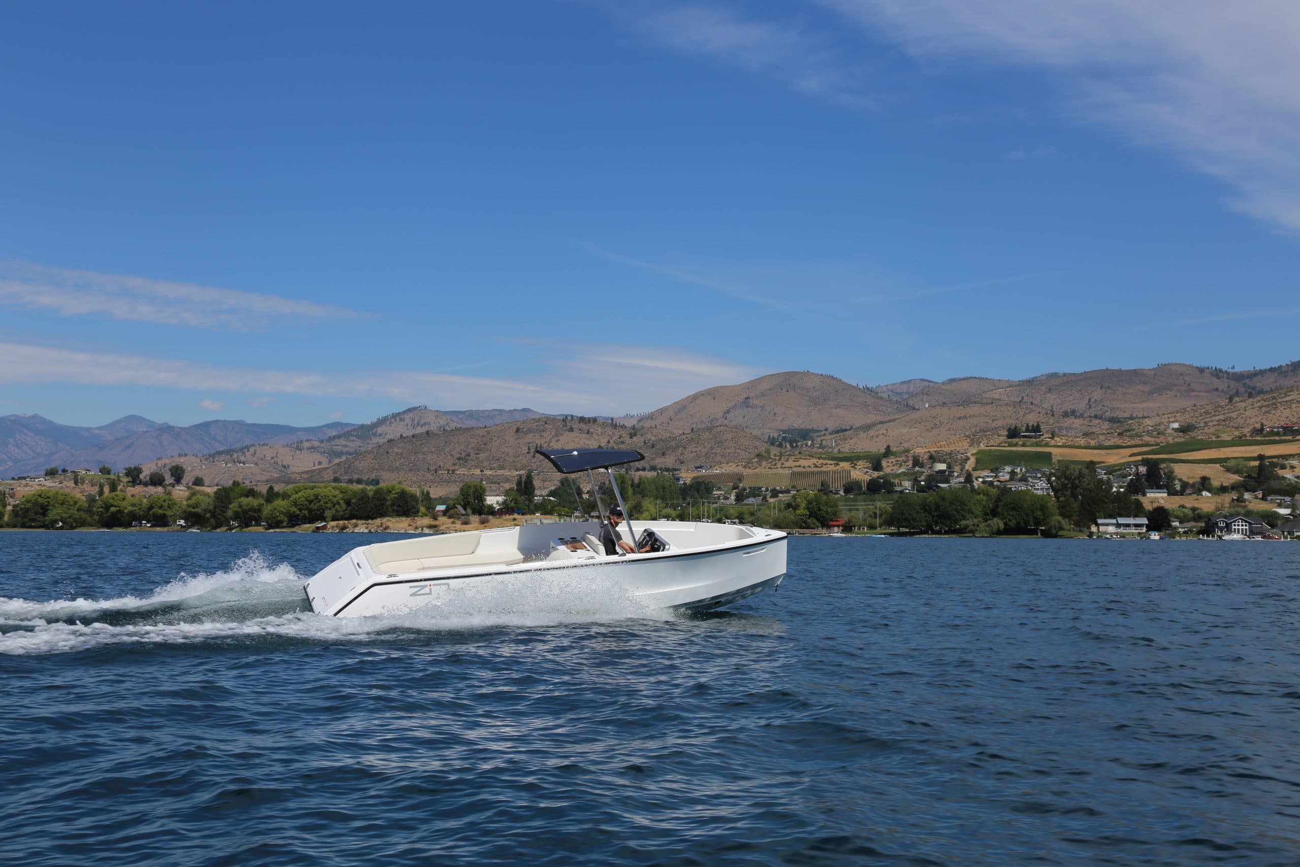 An electric boat cruising across a lake with mountain scenery in the background, creating a smooth wake under clear blue skies.