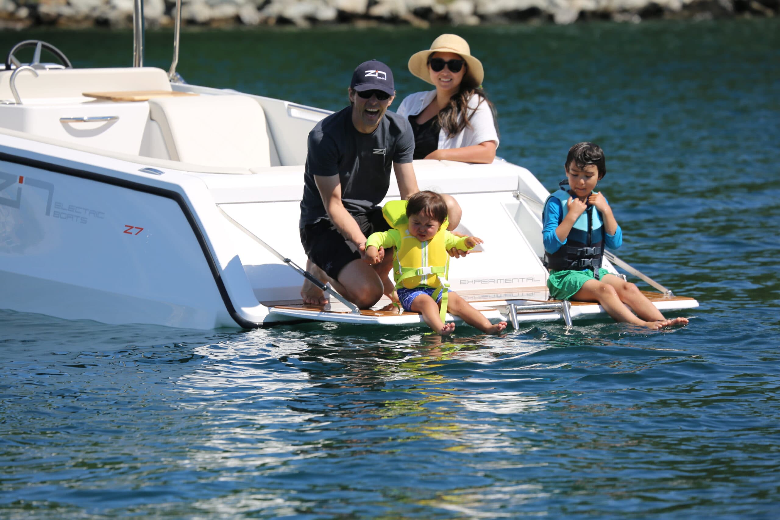 A family is sitting on the end of a boat (2026 Zin Boats Z7) there are two kids on the swim platform and one adult the other adult is wearing a hat and is sitting on the back of the boat.