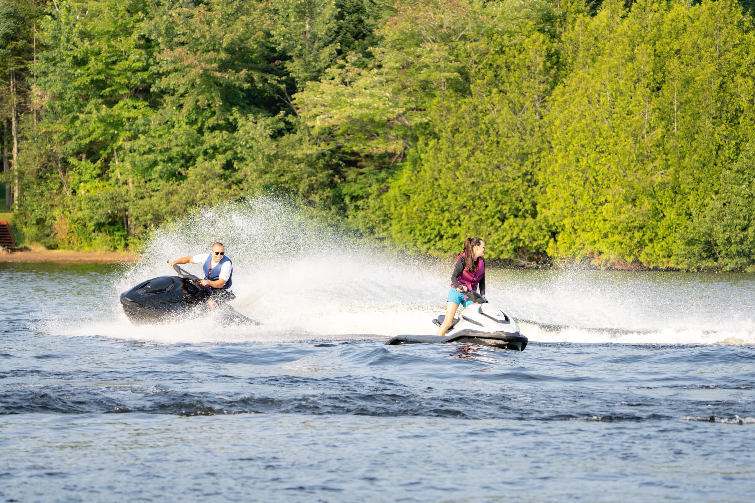 Two people riding personal watercraft (Taiga Orca P2) across a lake, viewed from shore with trees in the background.
