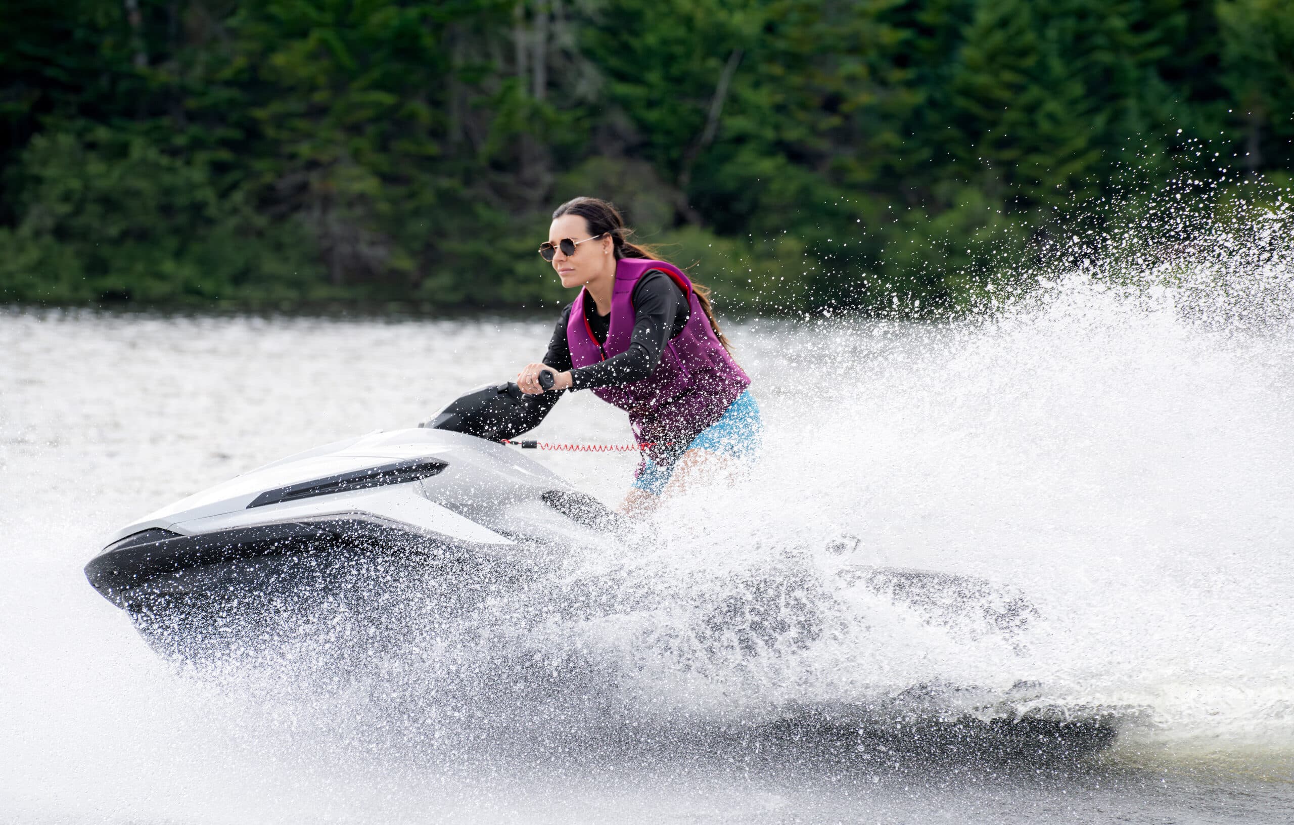 Rider operating a Taiga Orca Performance electric personal watercraft across a lake, creating a large spray with a forested shoreline behind.
