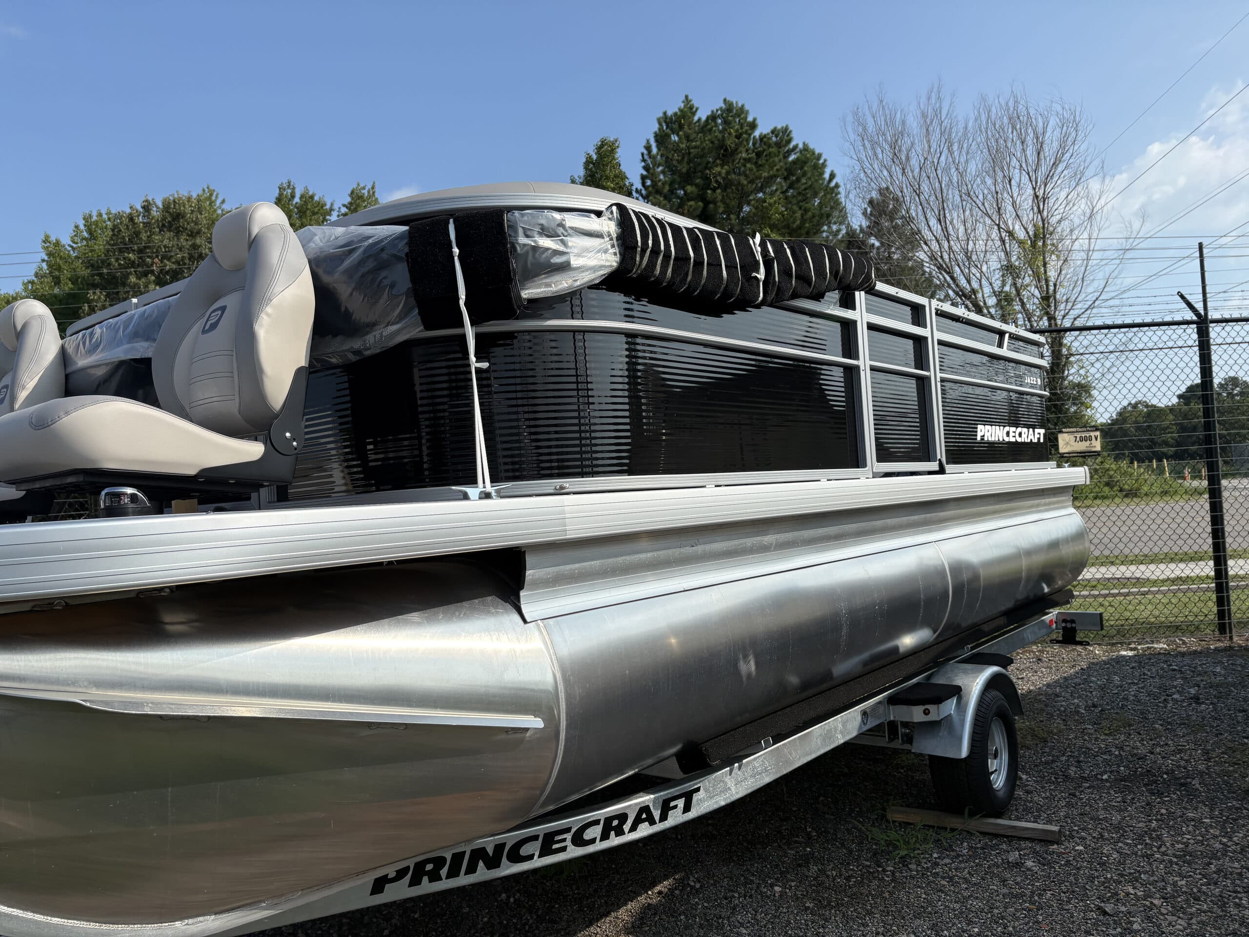 Side view of a Princecraft pontoon boat (2026 Princecraft Jazz 19 4s) on a trailer, showing the bow, side pontoons, railing, canopy, and seating.