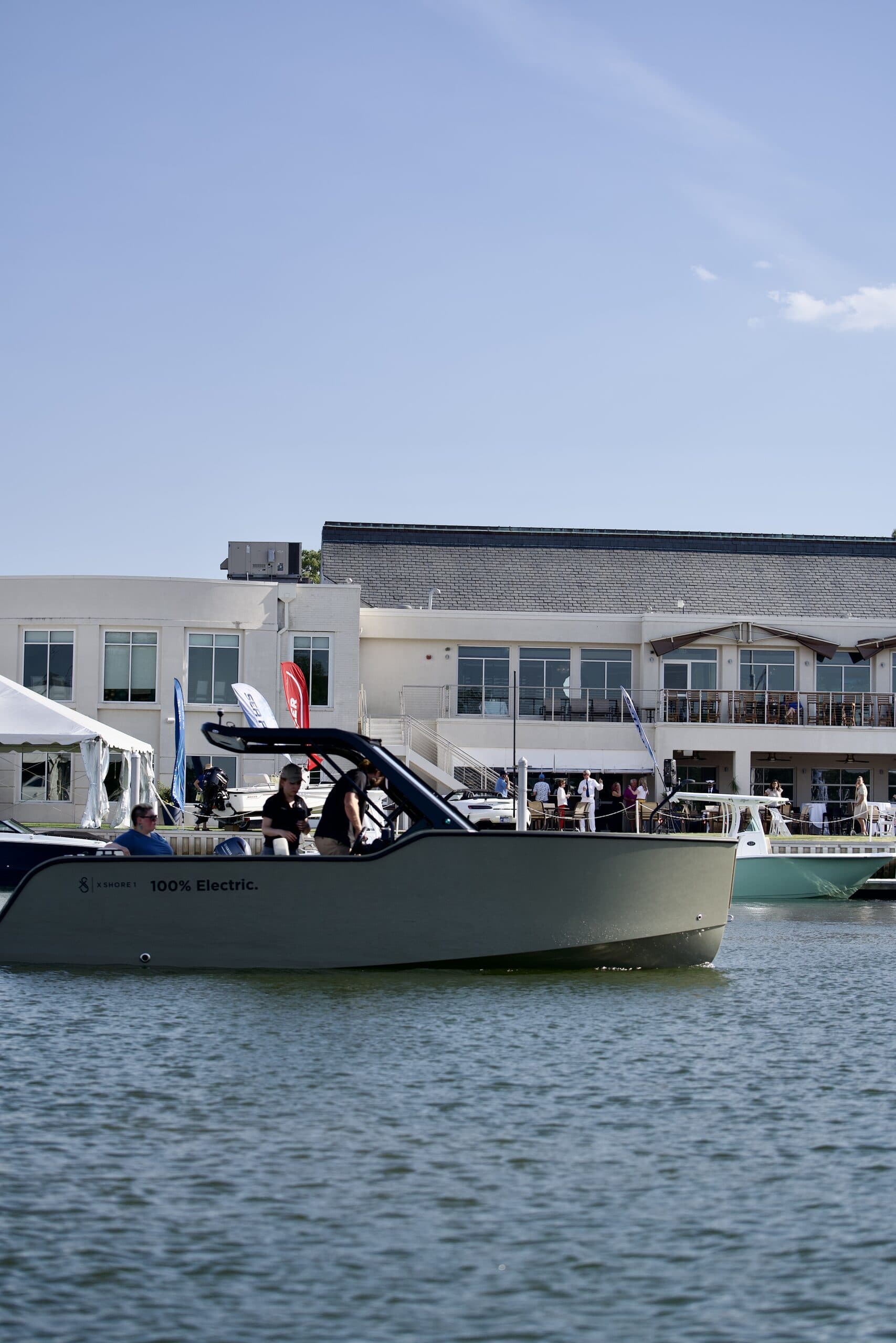 Side view of a X Shore 1 on the water, with passengers beneath a black canopy and waterfront buildings in the background.