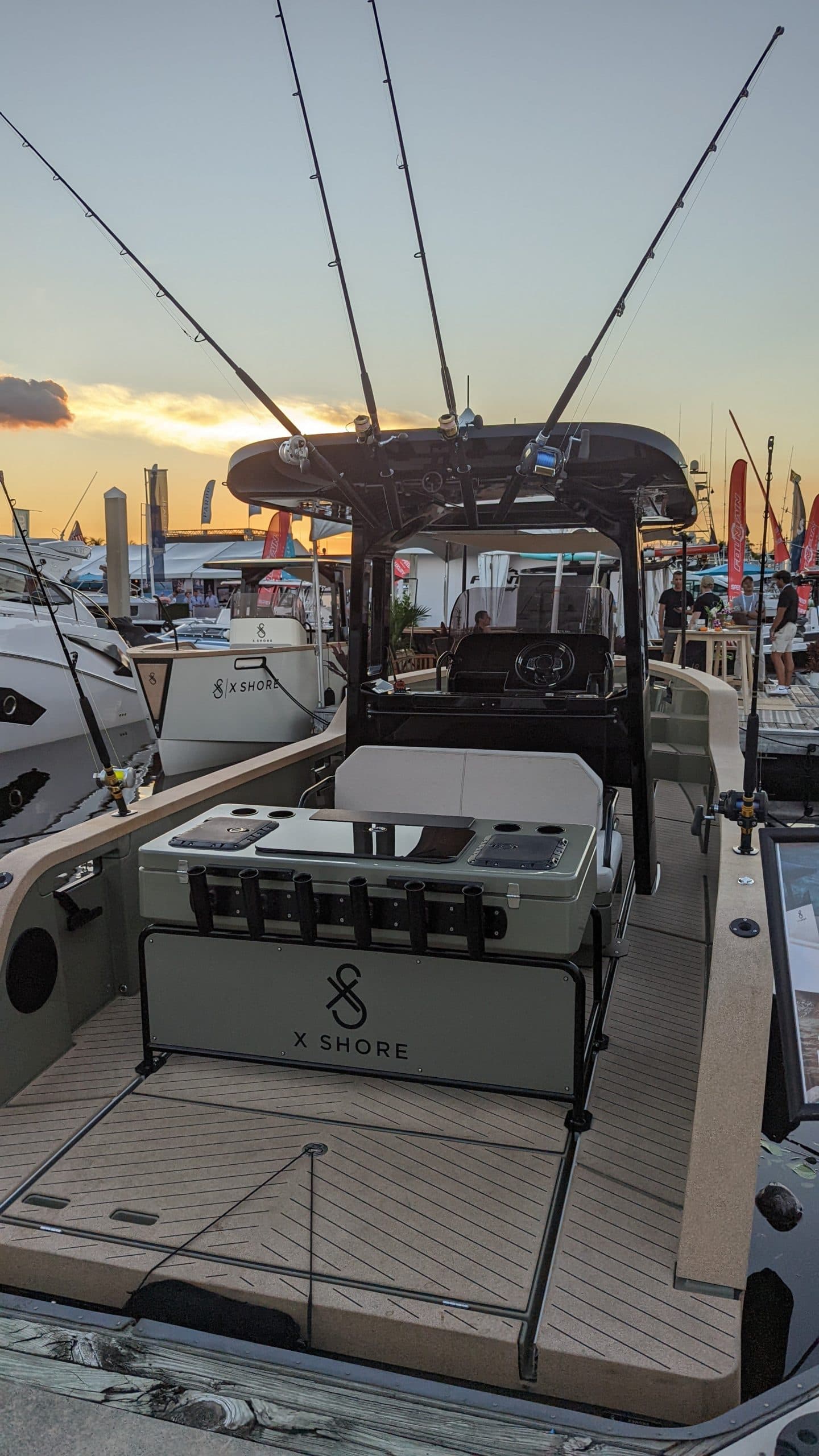 X Shore Eelex viewed from the stern, showing the aft deck, seating, helm, and roof-mounted fishing rods at a marina