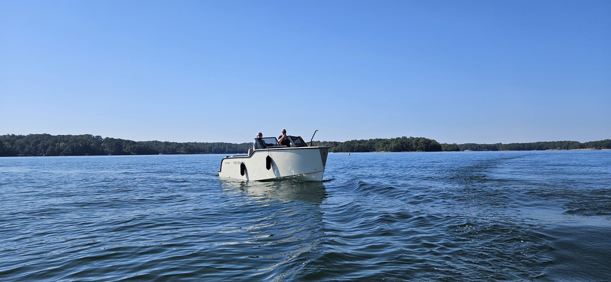 X Shore 1 viewed from the water at a distance, cruising on a lake with two people aboard and a wooded shoreline behind it.