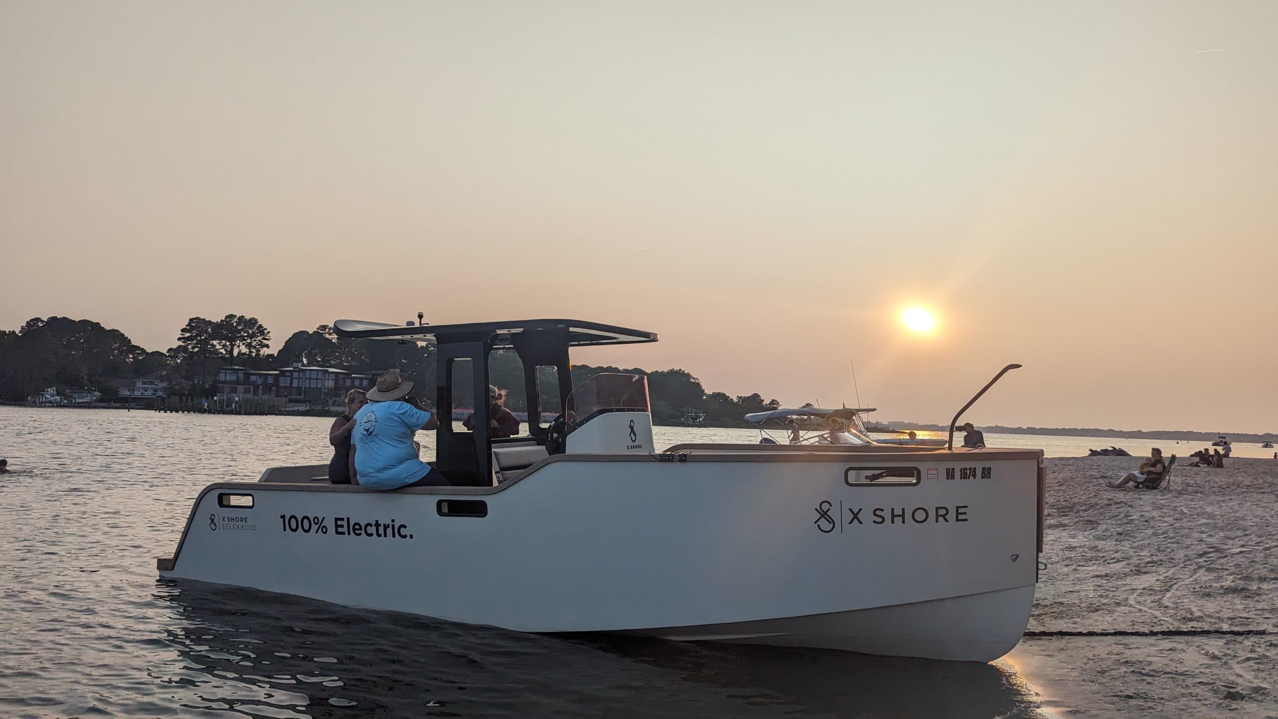 X Shore Eelex boat viewed from the port side on the water at sunset, with a person seated near the helm and “100% Electric” visible.