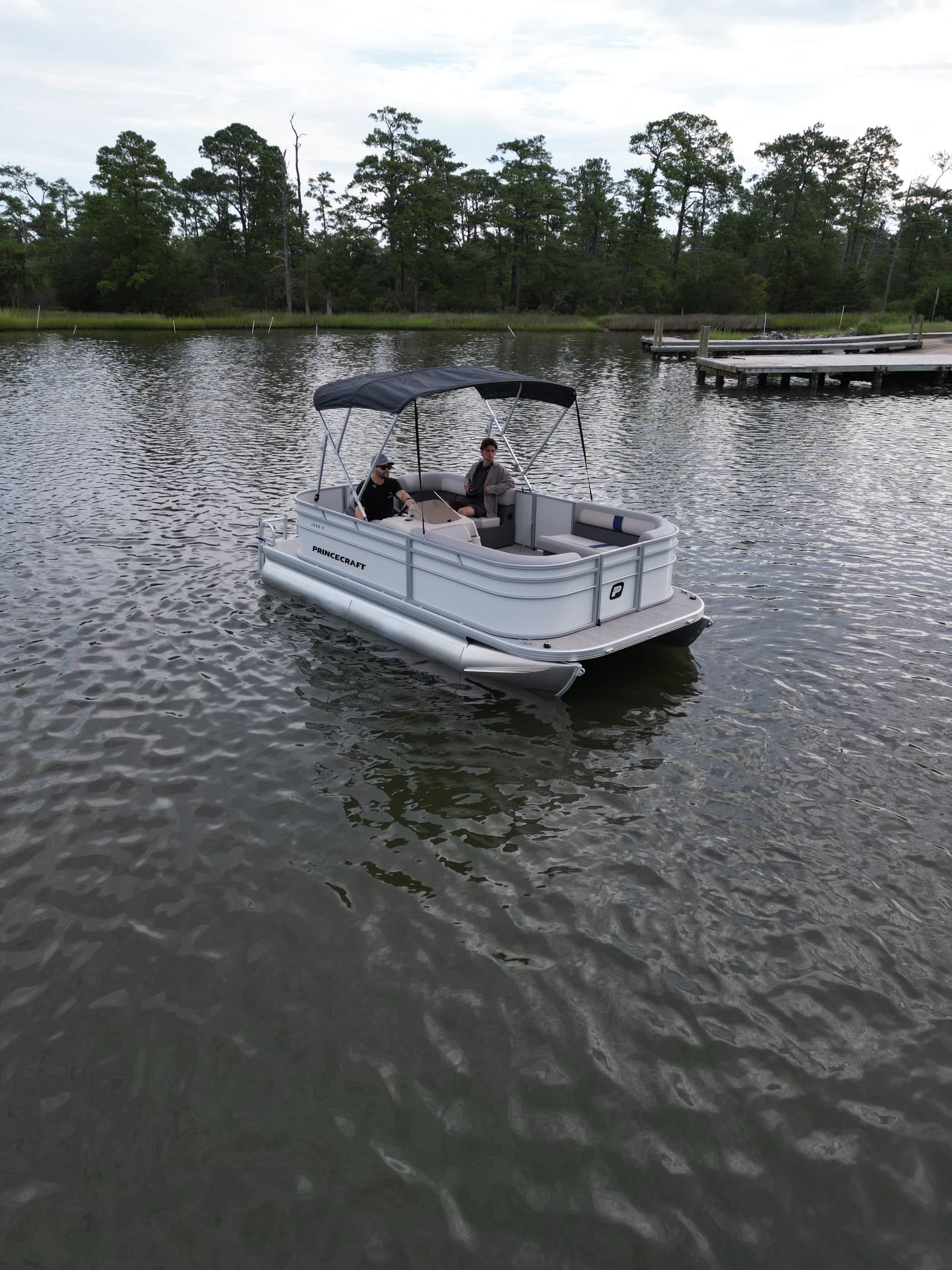 Princecraft Jazz 17 pontoon boat viewed from the rear three-quarter angle on a lake, with a canopy and two passengers aboard.