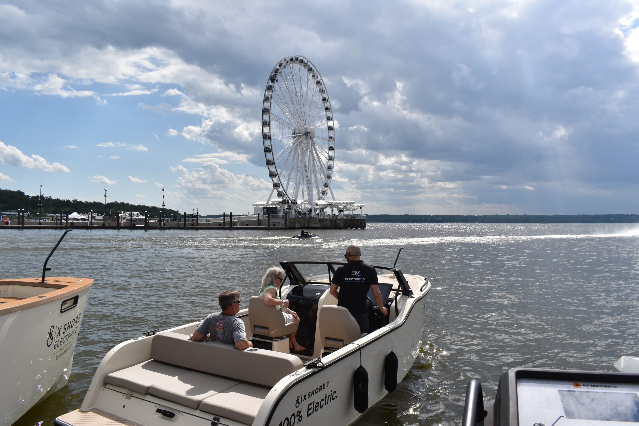 X Shore XS1 boat with people aboard, viewed from the stern near shore; a Ferris wheel and waterfront are visible in the background