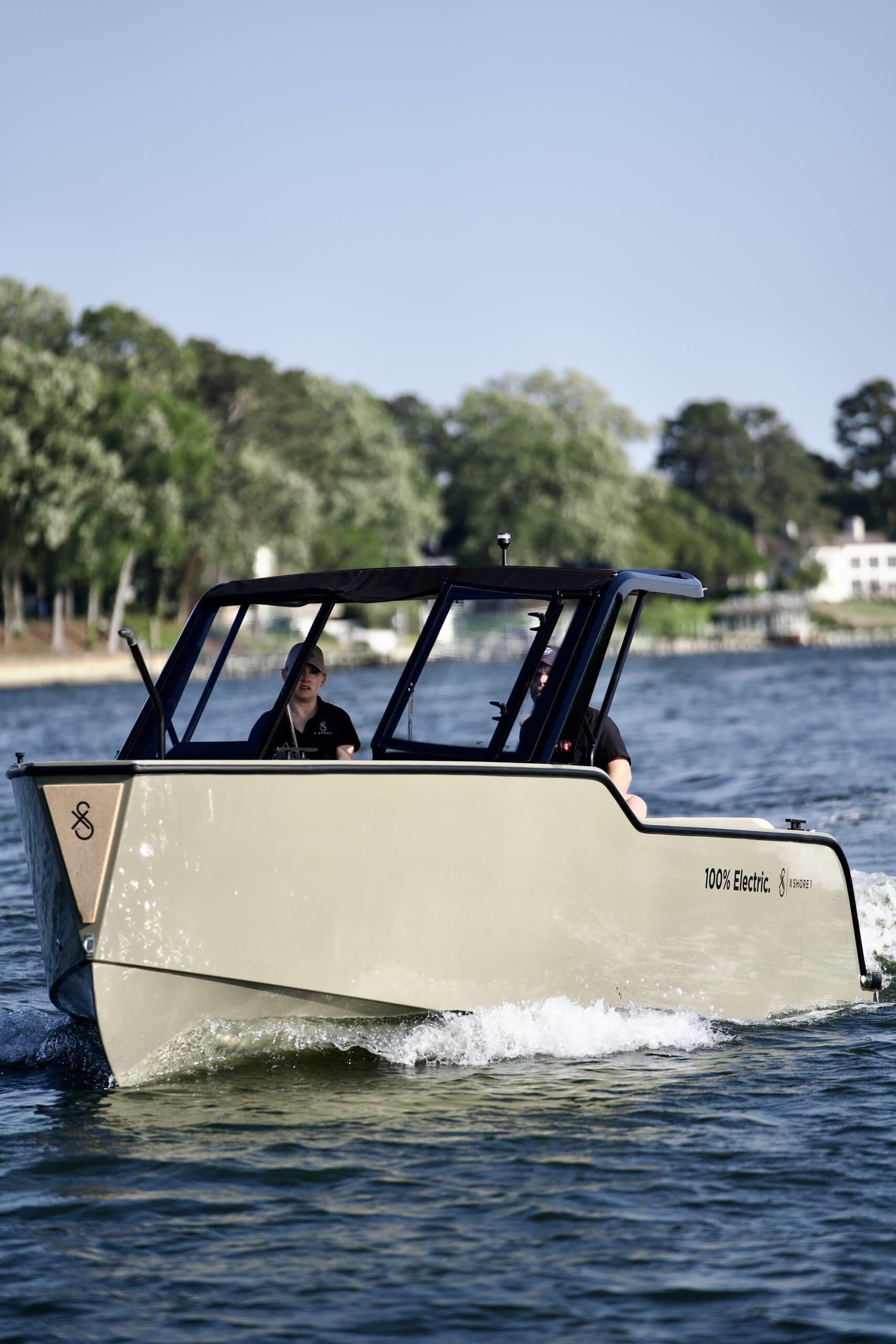 Side view of a cream-colored X Shore cruising on a river, with a dark canopy and windshield visible.