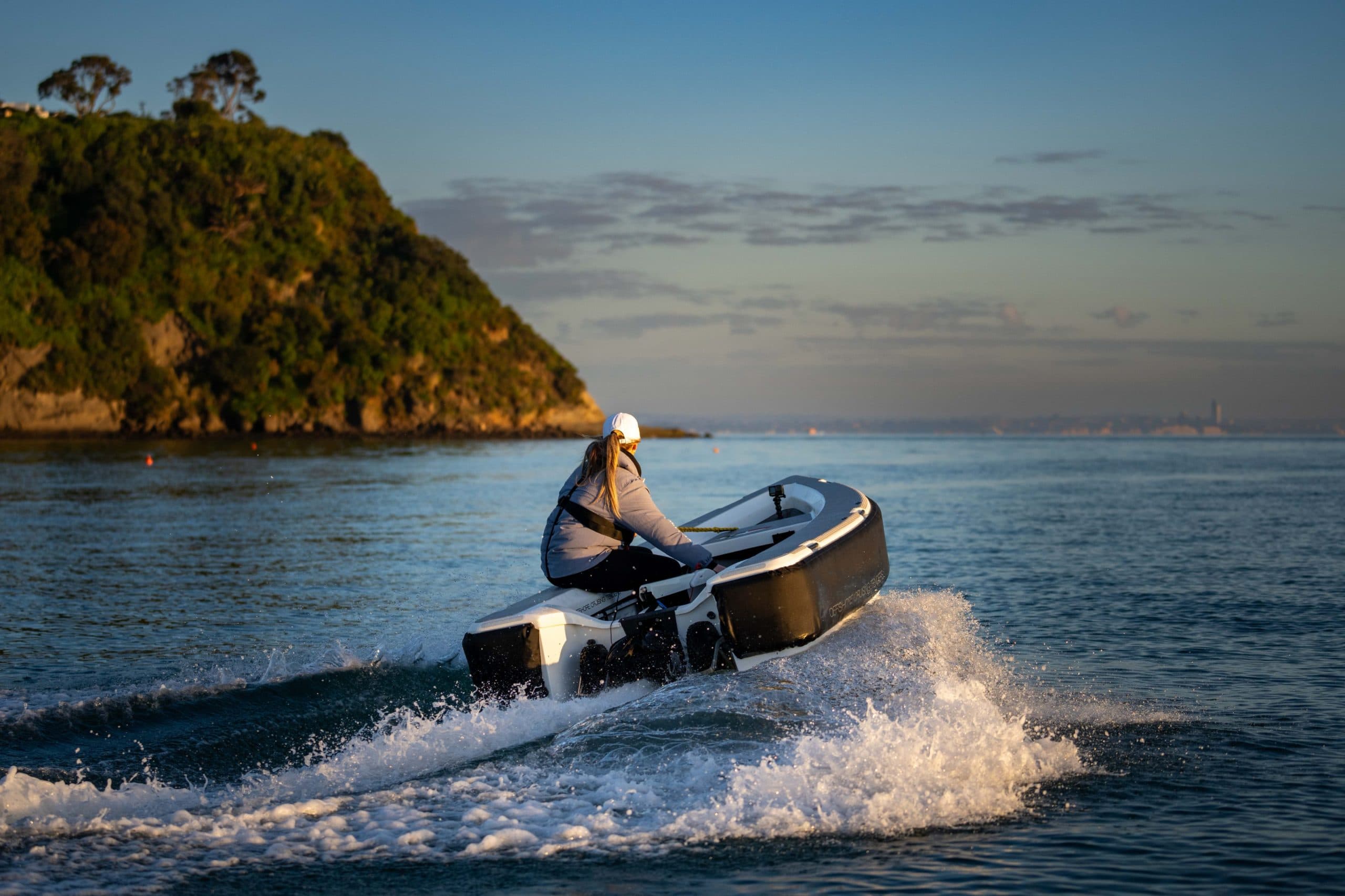 Offshore Cruising Tenders OC330 speeding across coastal water, viewed from the side with a person at the helm and shoreline behind.