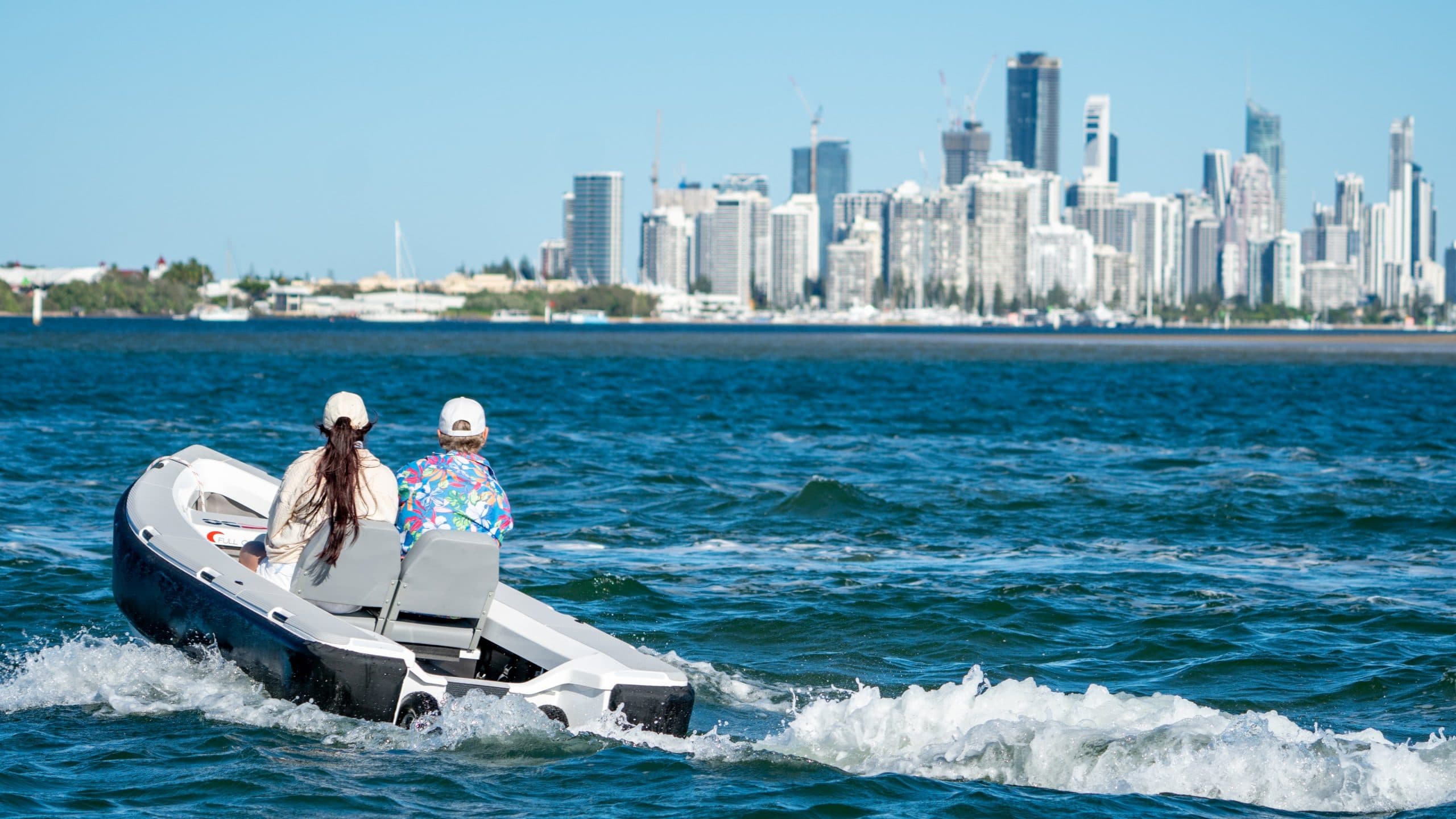 Small 2026 Offshore Cruising Tenders OC350 speeding across the water, viewed from behind, with two passengers and a city skyline in the background.