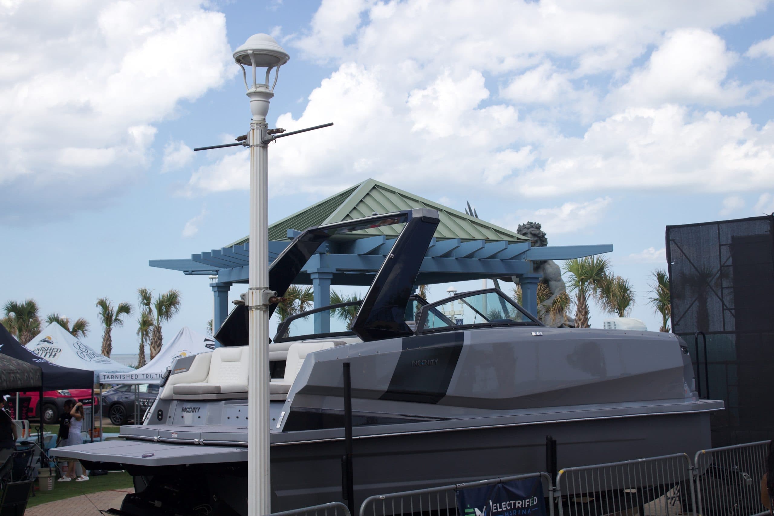Side view of a gray wake boat displayed at a marine event, with its tower raised and cockpit partially visible.