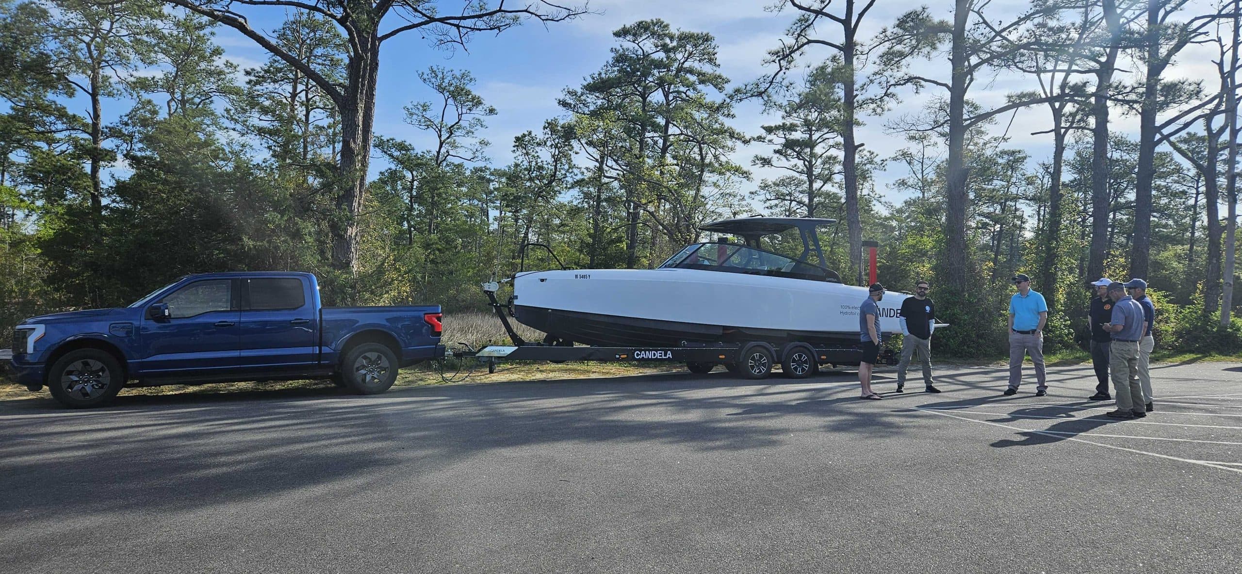 Candela C-8 on a trailer beside a pickup truck, viewed from the side in a wooded parking area.