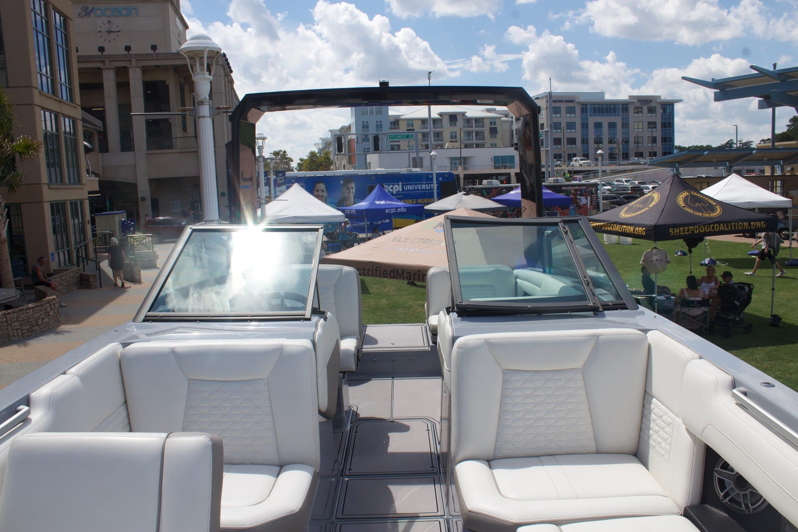 View from the bow into the cockpit of a white wake boat, showing the windshield, helm, seating, and tower at a waterfront event.