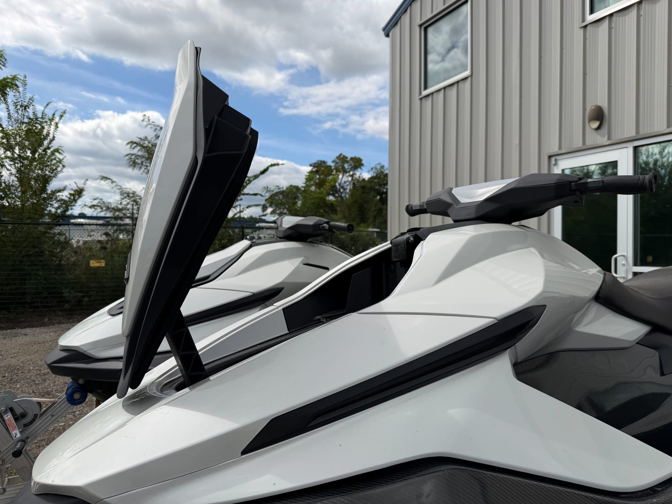 Close view of two white personal watercraft beside a building, with the front craft’s raised hatch and handlebars visible.