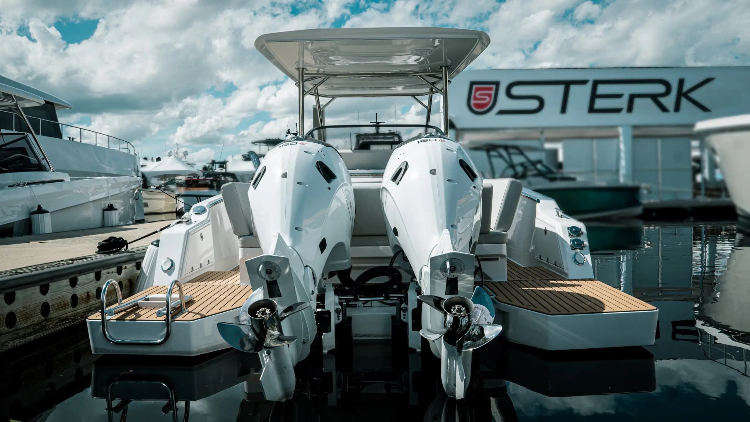 Rear view of a white Sterk 31E-style motorboat with twin outboard motors at a marina, with a Sterk sign in the background