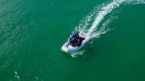 Aerial view of a small rigid inflatable boat underway, leaving a curved white wake on green water.