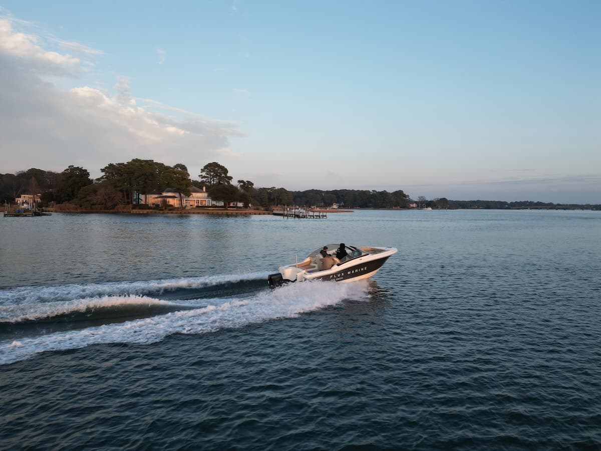 Scout 215 Dorado motorboat moving at planing speeds across calm coastal water, leaving a wake near a tree-lined shoreline.