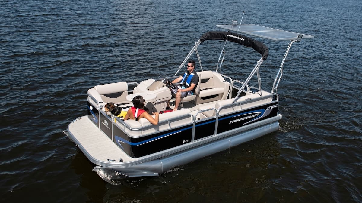 Three-quarter elevated view of a Princecraft Brio e 17 pontoon boat underway, showing the bow, cockpit seating, canopy, and two occupants.