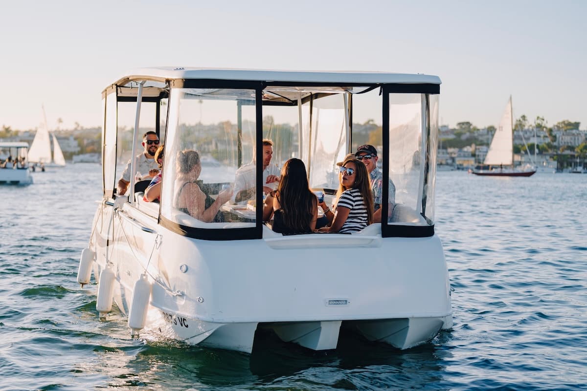 ElectraCraft 182TR electric boat carrying passengers on calm water, with clear canopy, nearby sailboats, and waterfront homes in the background.