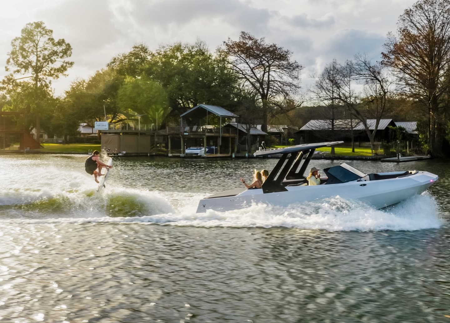 White center-console boat (2026 Voltaic AEW24) towing a wakeboarder across a lake near a marina, with passengers aboard and spray trailing behind.