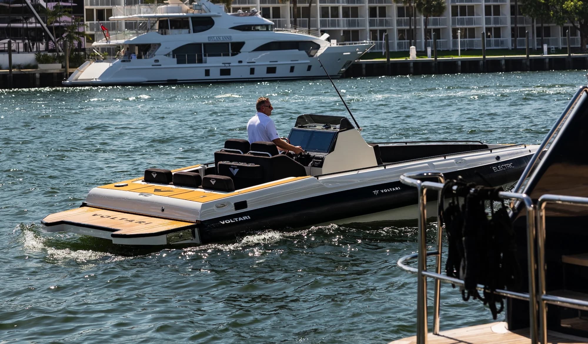 Voltari electric boat cruising on a sunny waterfront, with a seated operator, open deck, and yacht-filled marina behind it.