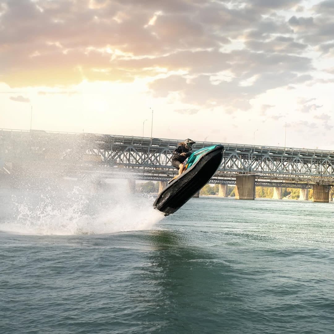 Taiga Orca Performance electric personal watercraft jumping above the water, with a bridge and sunset sky in the background.