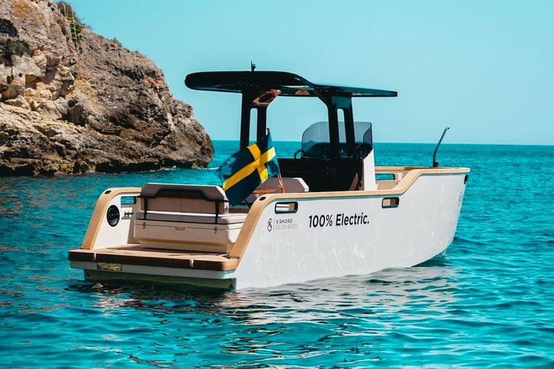 X Shore Eelex boat viewed from the port rear quarter, showing the stern, open cockpit, console, and rocky coastline.