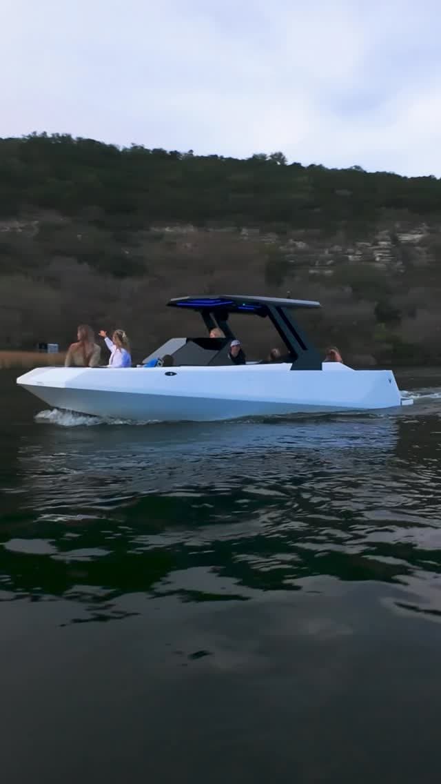 Side view of a white recreational motorboat with a dark canopy, passengers aboard, and a wooded rocky shoreline in the background (2026 Voltaic AEW24).