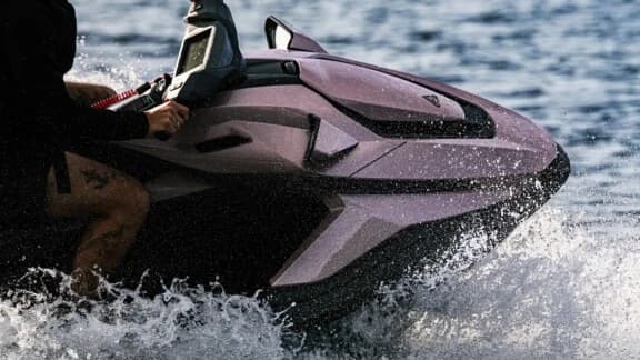 Close-up side view of a pink personal watercraft (2026 Taiga Orca WX3) moving across choppy water, showing the bow and rider’s hands at the handlebars