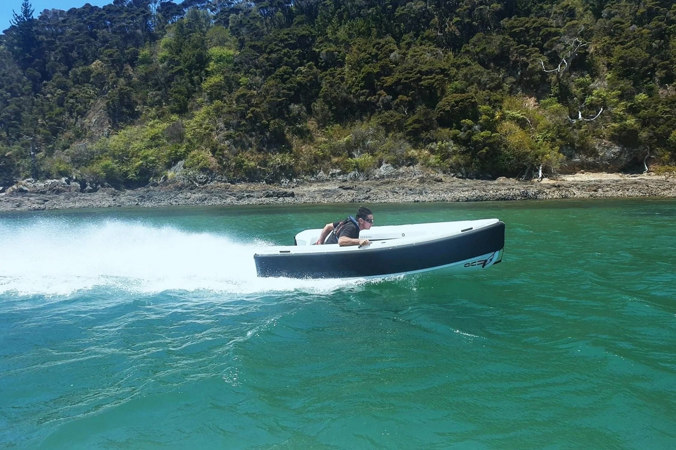 Small black-and-white 2026 Offshore Cruising Tenders OC330 speeding across turquoise water near a forested shoreline, viewed from the side.