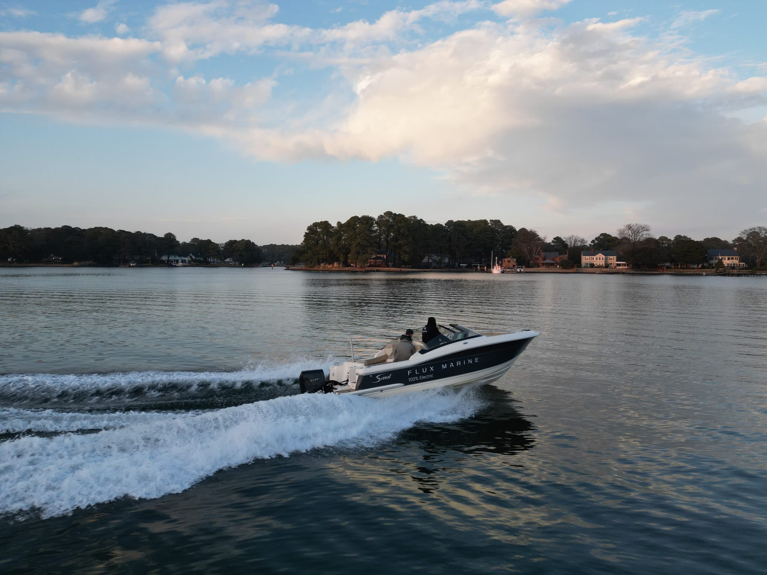 Scout 215 Dorado bowrider boat turning in calm water, leaving a foamy wake with a tree-lined shoreline in the background.