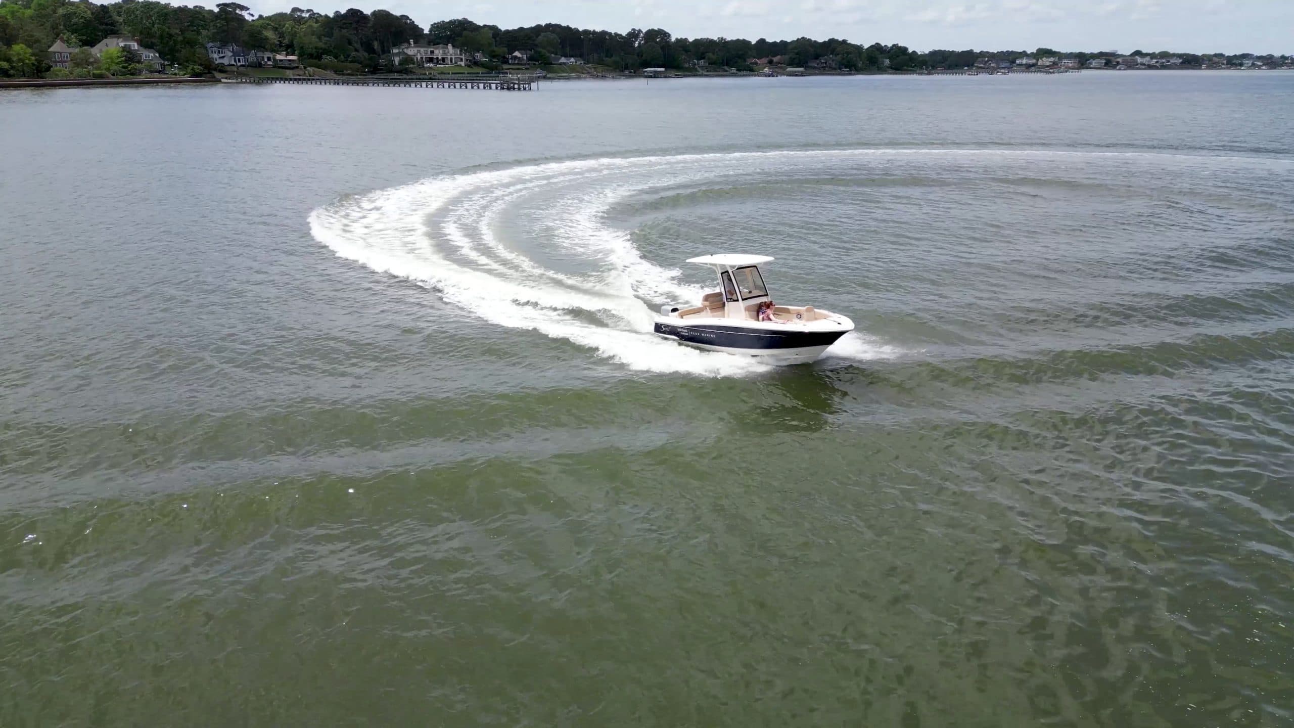 Electric Scout 215 XSF motorboat turning across calm bay water, leaving a curved white wake with a tree-lined shoreline in the background.