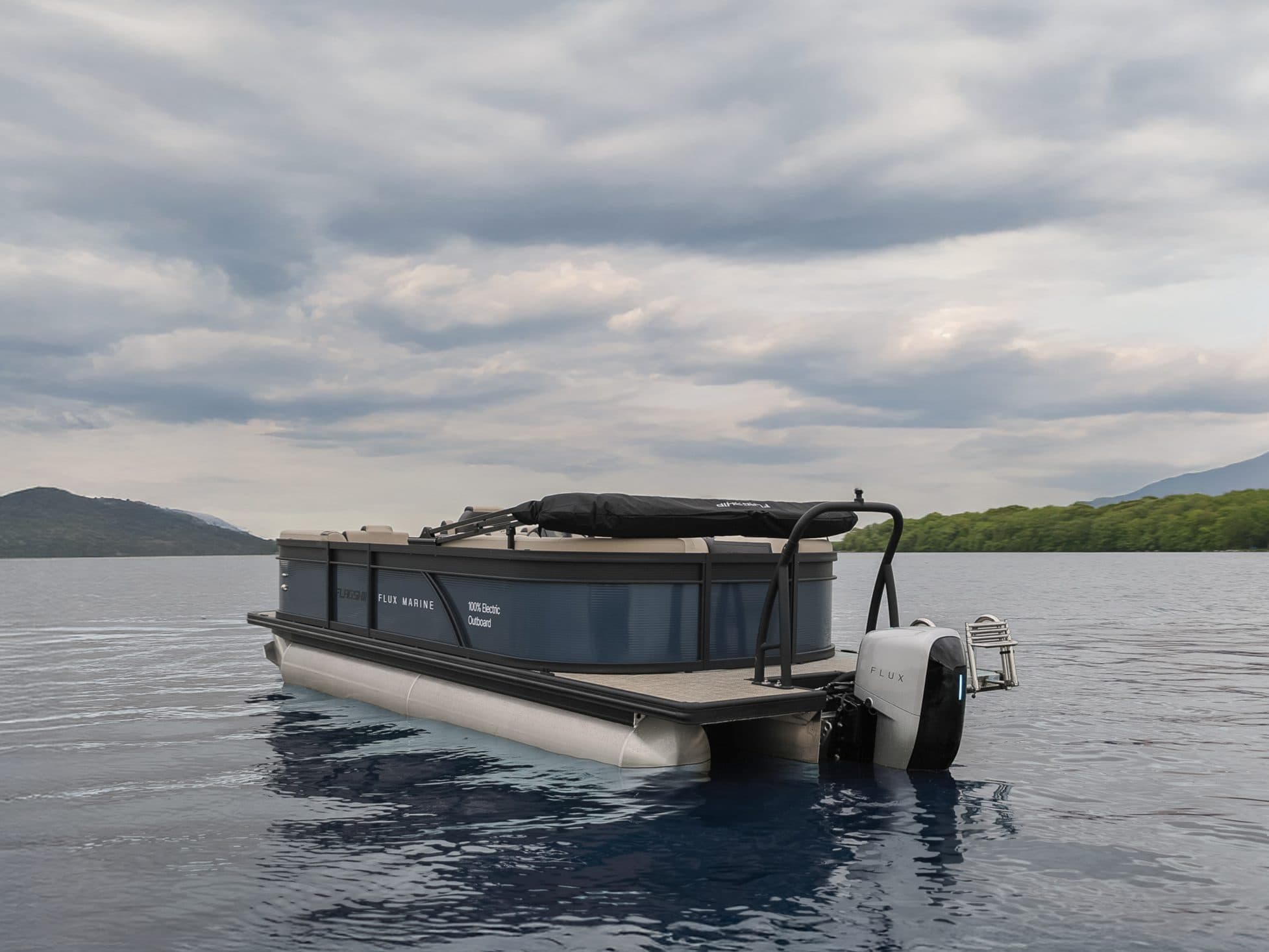 Flux Marine Discover pontoon boat with an outboard motor on calm water, with forested hills and a cloudy sky in the background.