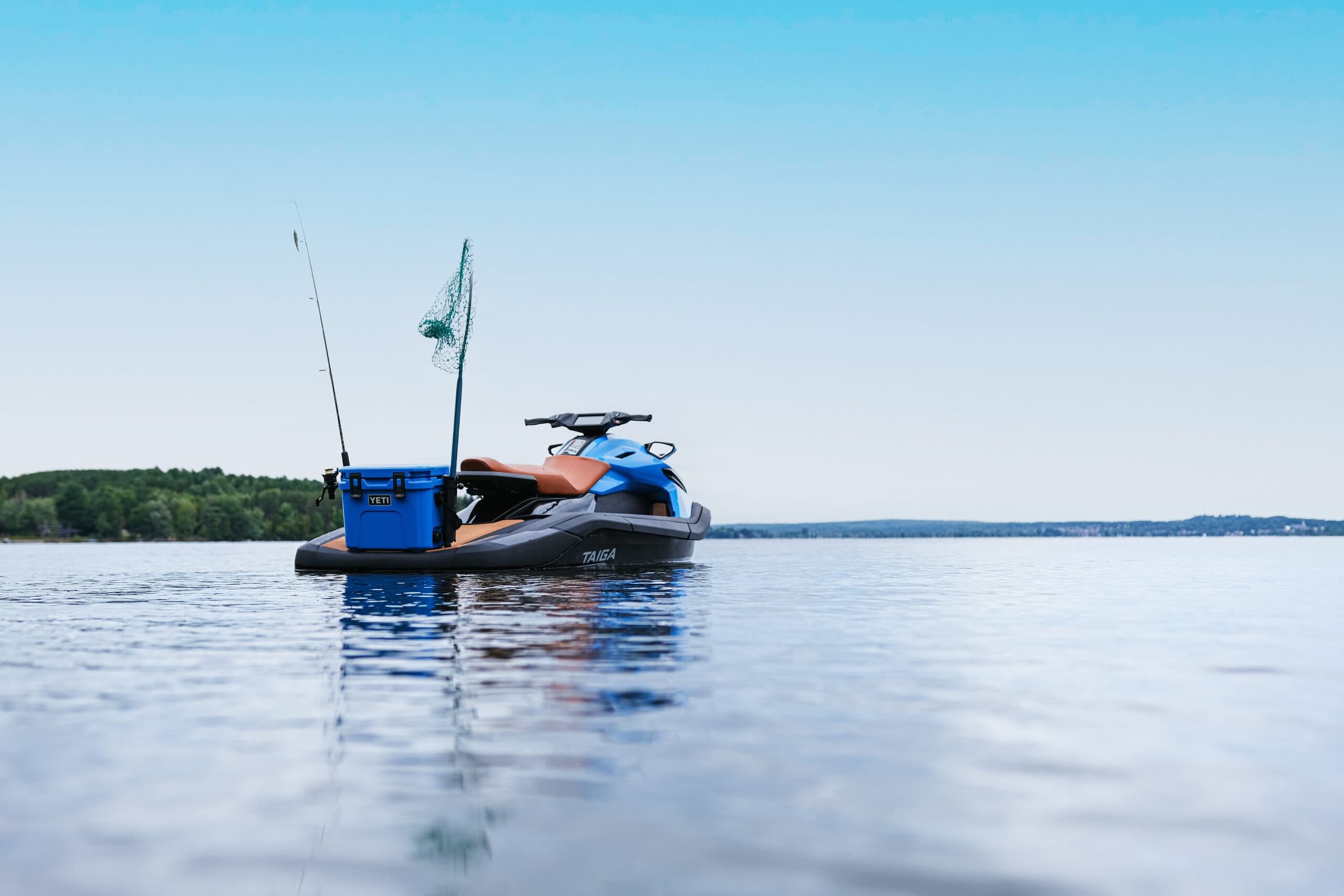 Taiga Orca WX3 personal watercraft viewed from the side on a calm lake, with a blue cooler and fishing rods visible.